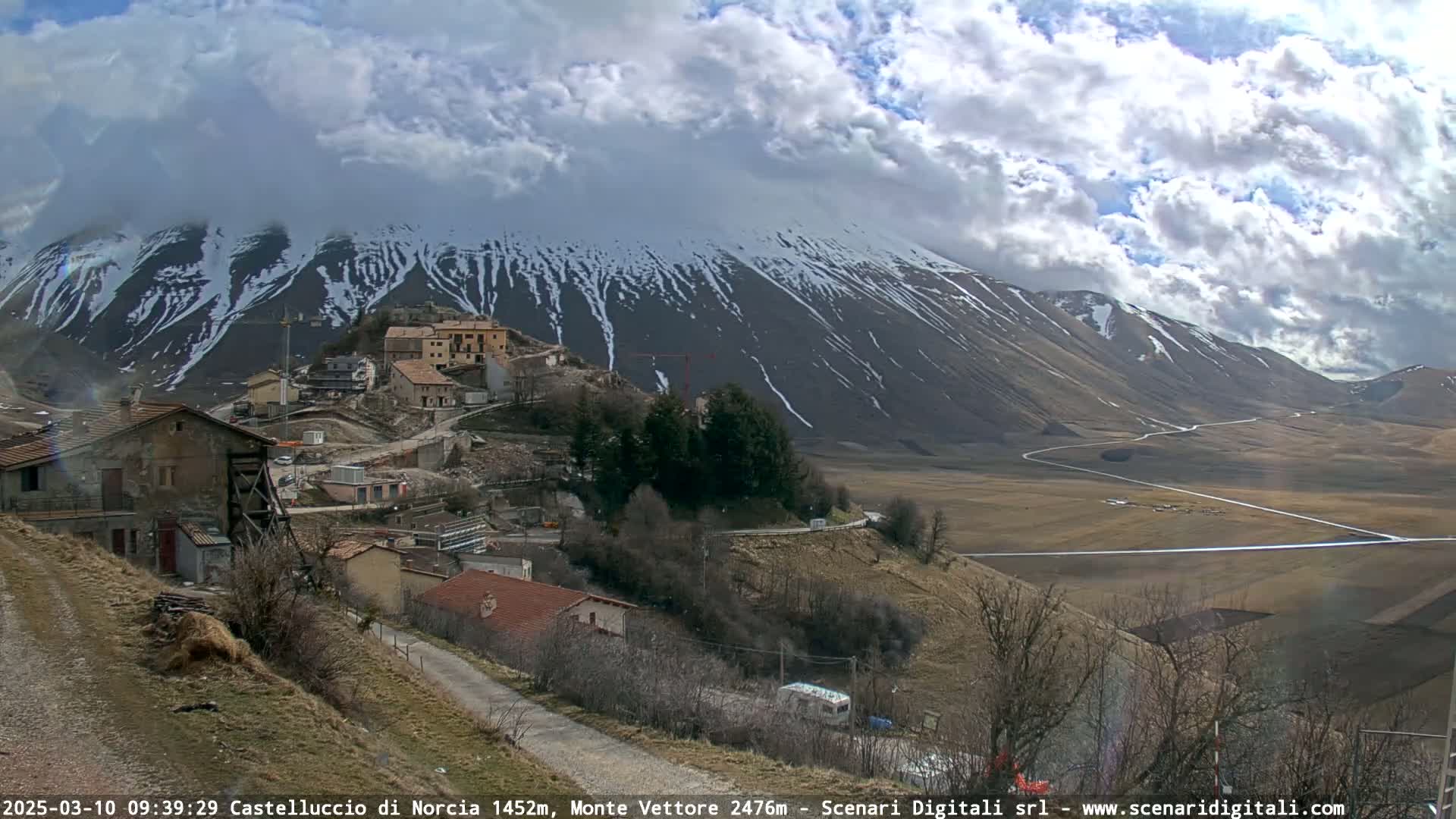 A partly cloudy day reveals a snow-dusted mountain range overlooking a small village nestled in a valley with winding roads.