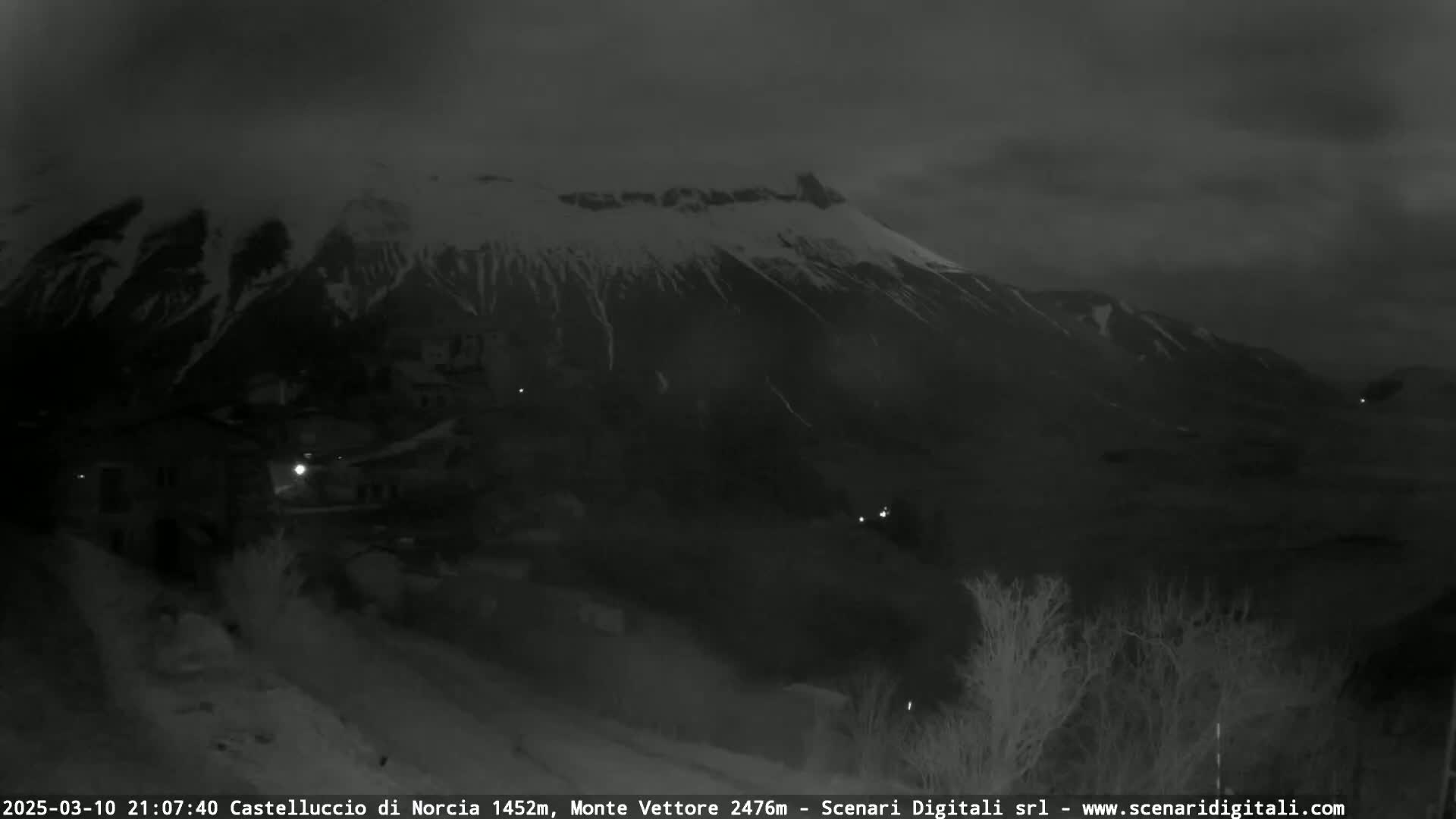 A nighttime, low-light view shows a snow-covered mountain range behind a small village with a few illuminated windows.