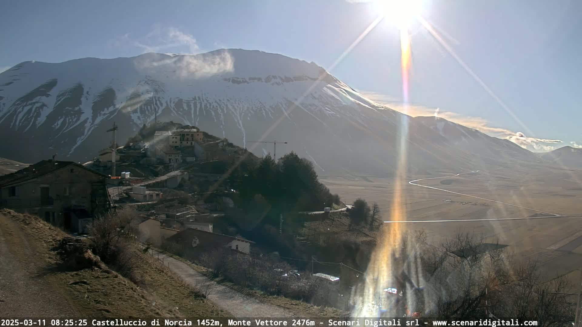 A sunlit village nestled in a valley below a snow-capped mountain range on a bright, clear day.
