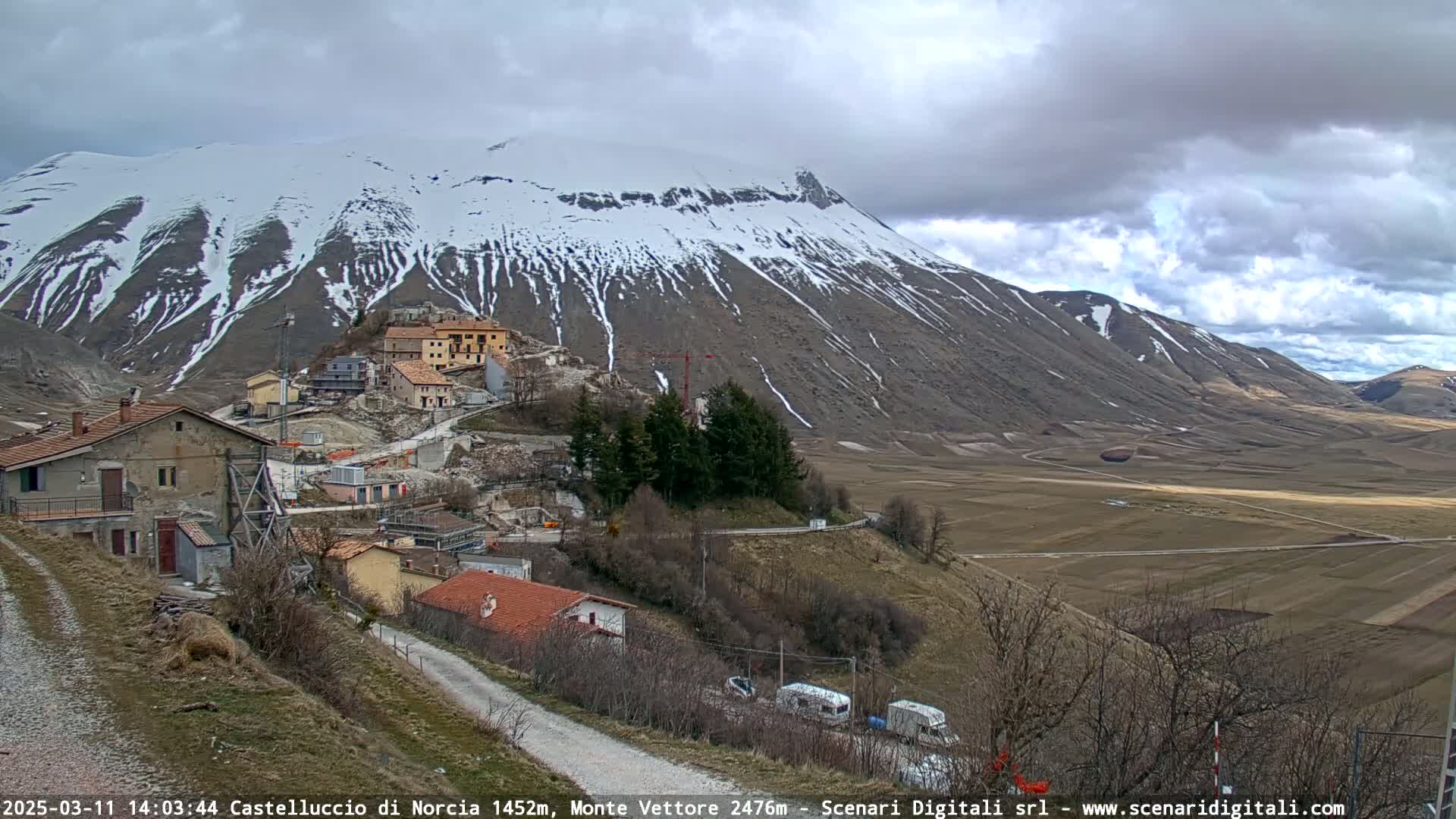 A snow-capped mountain overlooks a small village nestled in a valley under a cloudy sky.