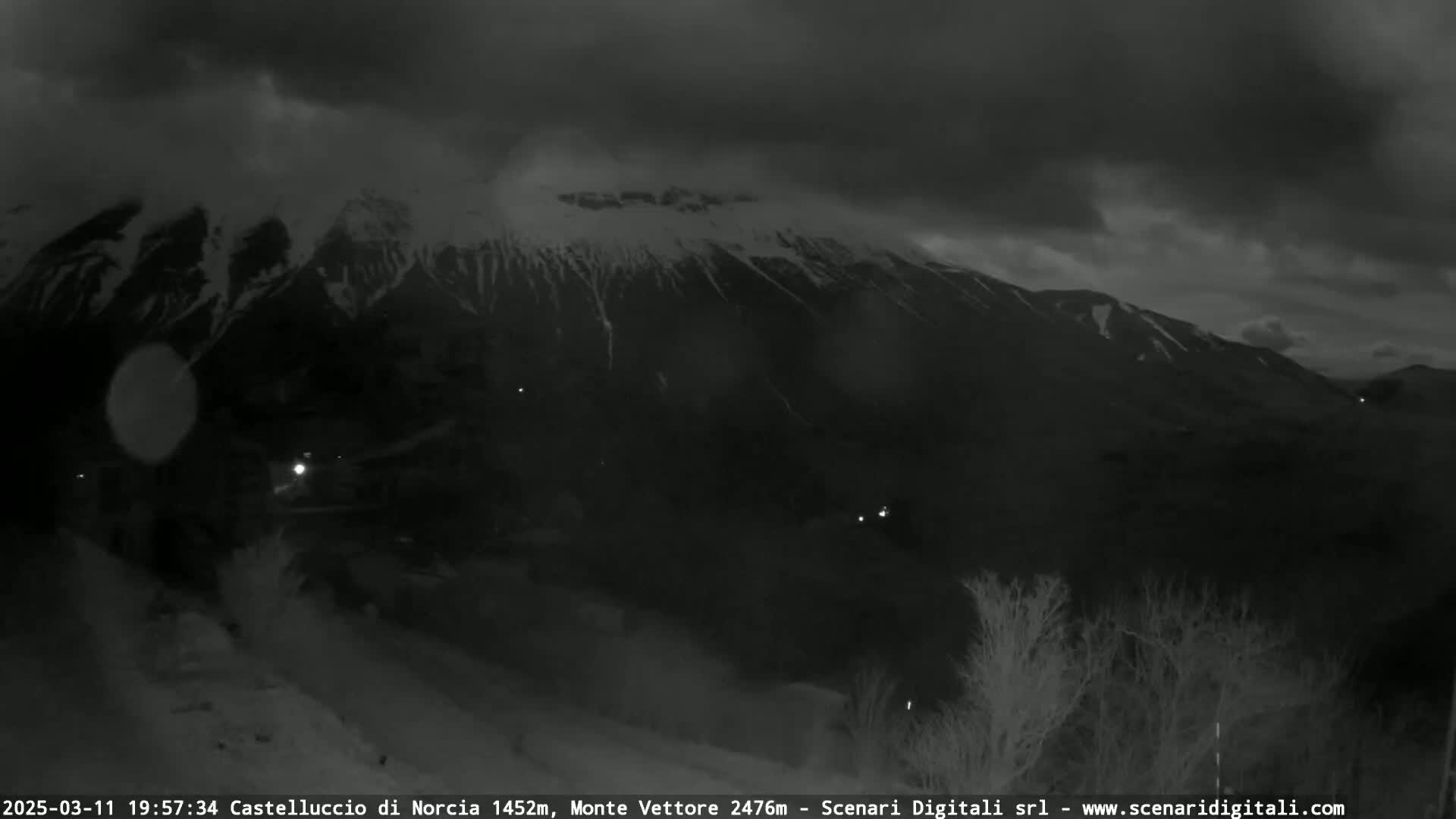 A nighttime, overcast view shows a snow-capped mountain range in the distance, with a few faint lights visible in the foreground.