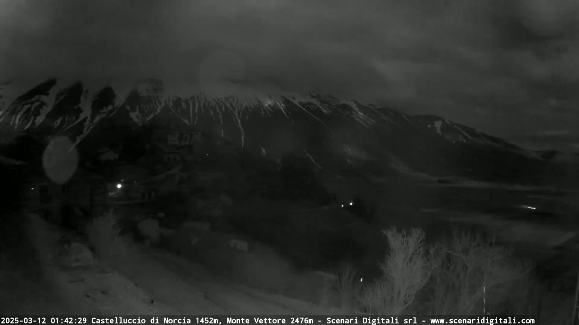 A nighttime, overcast view shows a snow-dusted mountain range behind a dimly lit town.