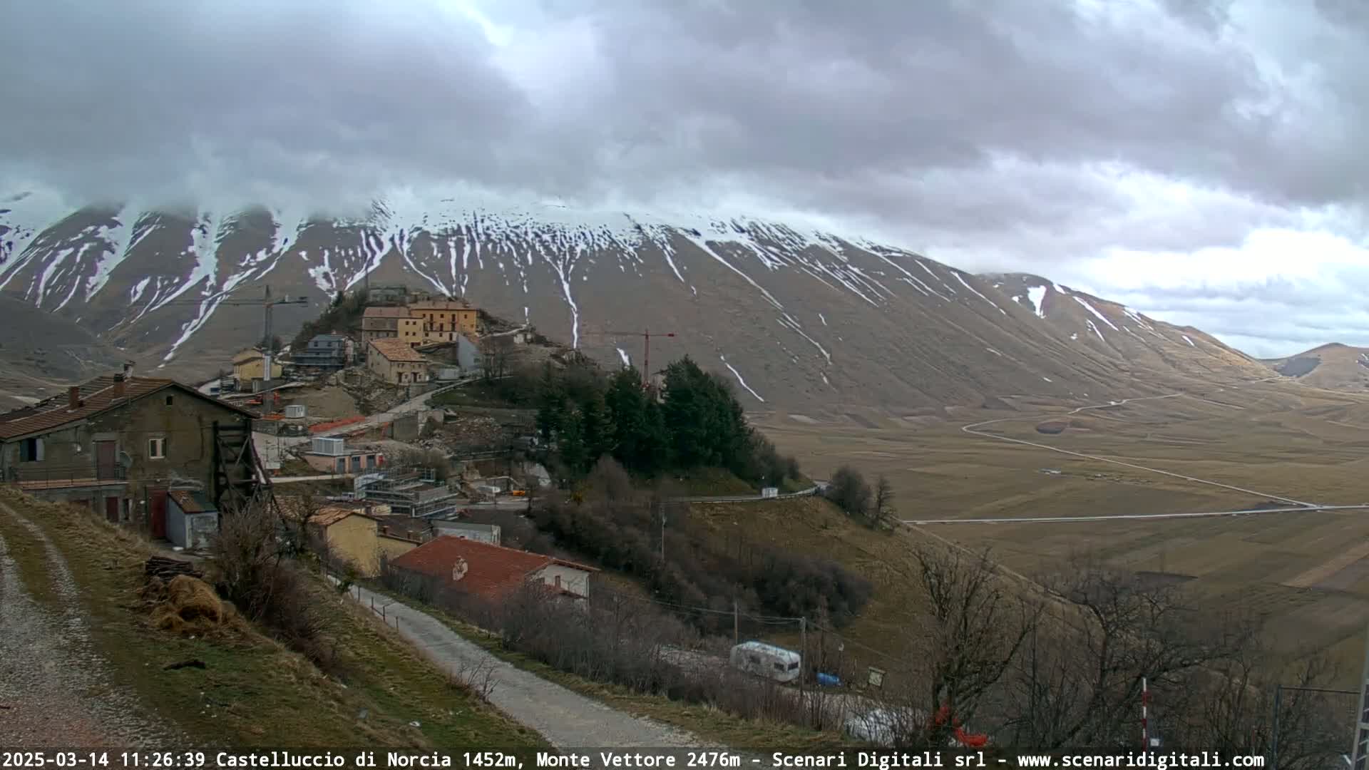 A small village nestled on a hillside beneath a snow-dusted mountain range on an overcast day.