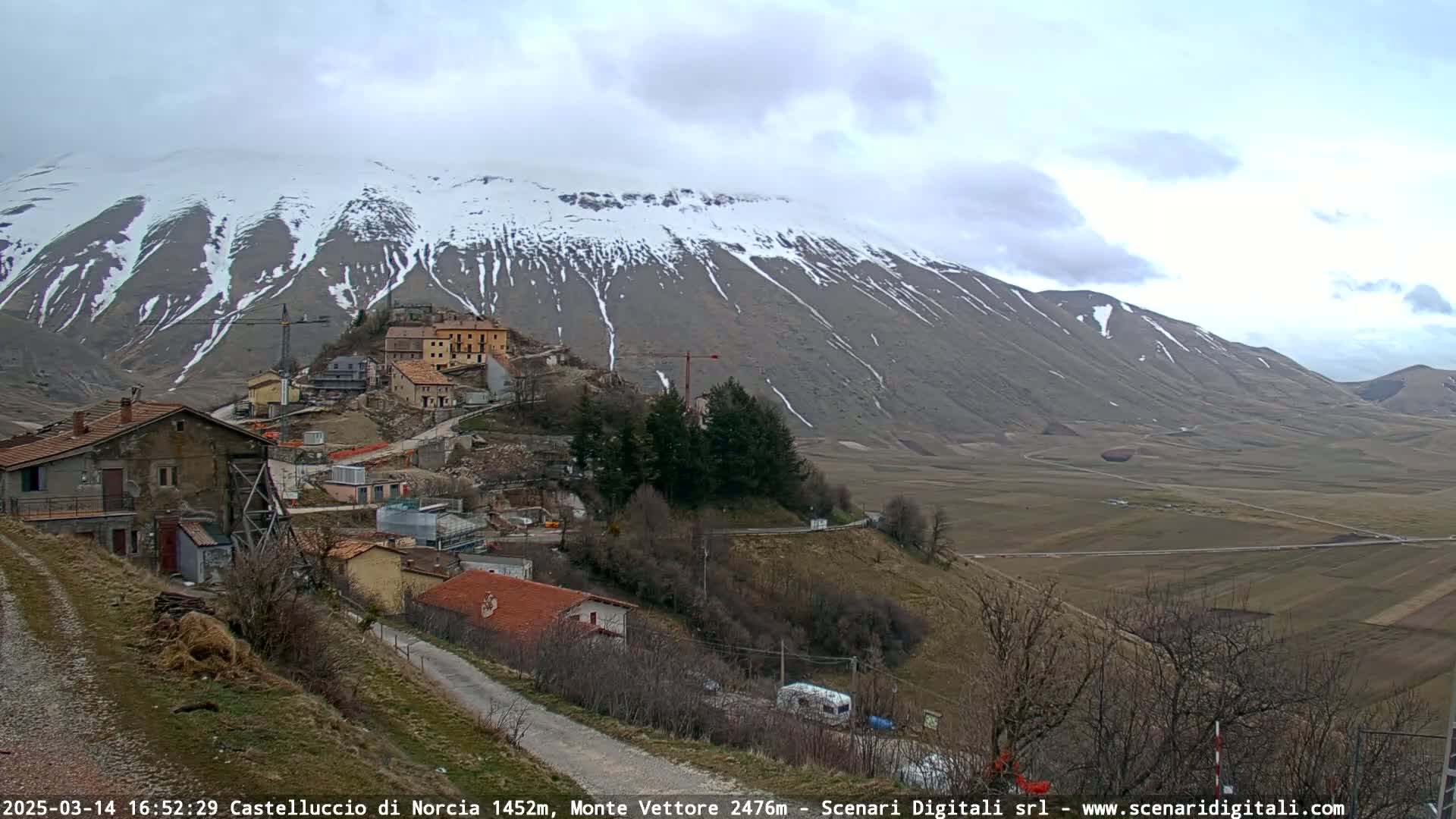 A partially snow-covered mountain overlooks a small village nestled in a valley under a cloudy sky.
