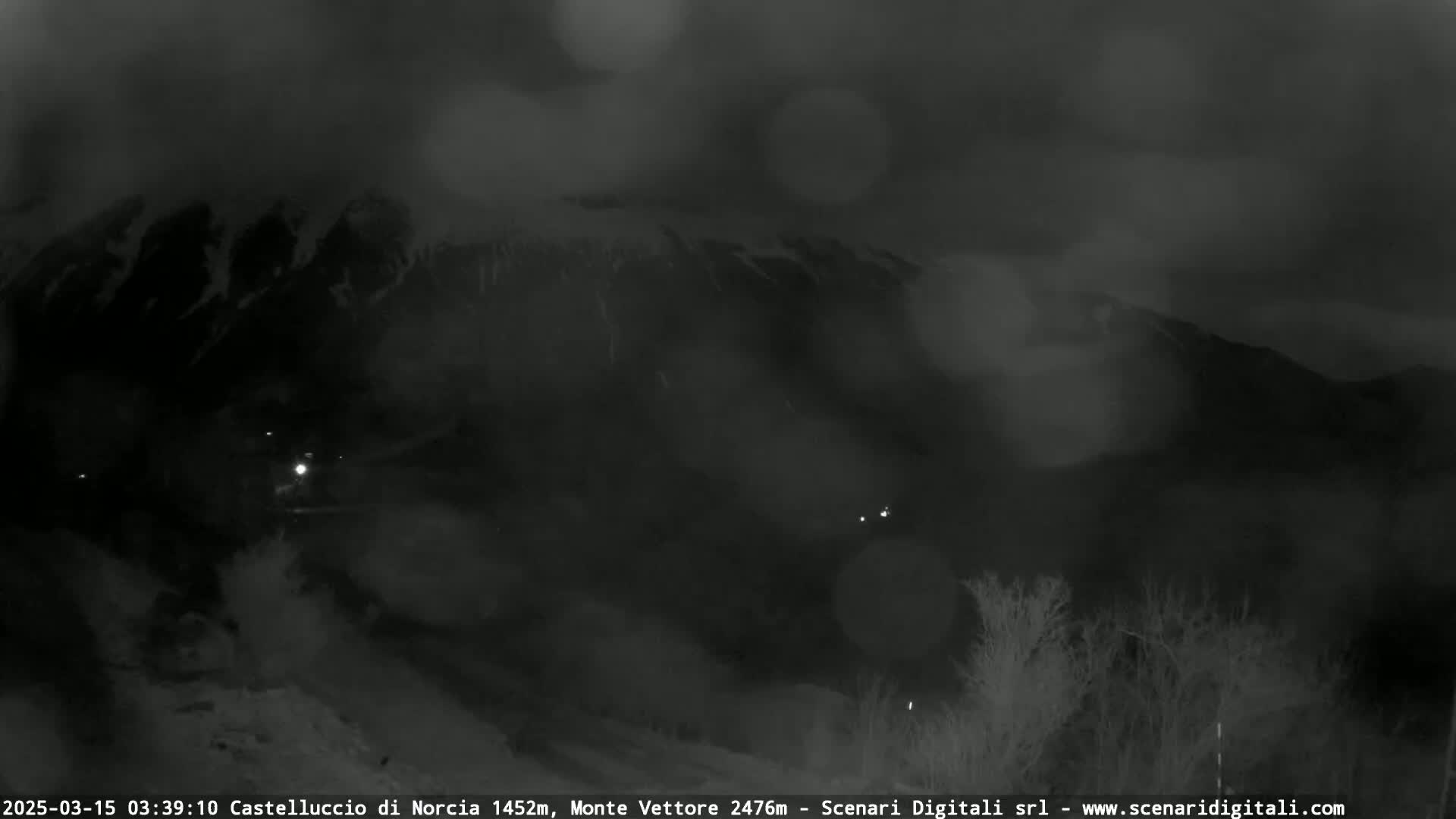 A nighttime, foggy view of a snow-covered mountain range with sparse vegetation in the foreground.