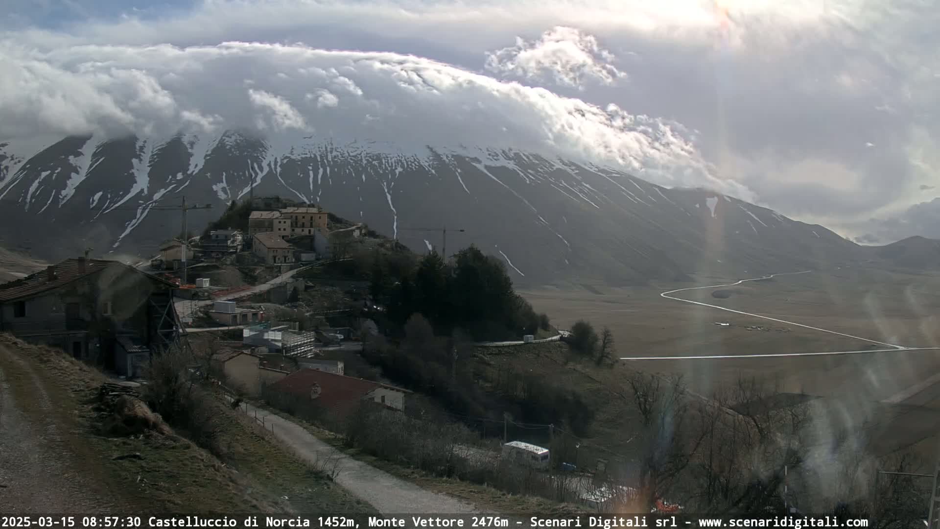 A partly cloudy day reveals a snow-dusted mountain range behind a small village nestled in a valley, with a winding road visible in the distance.