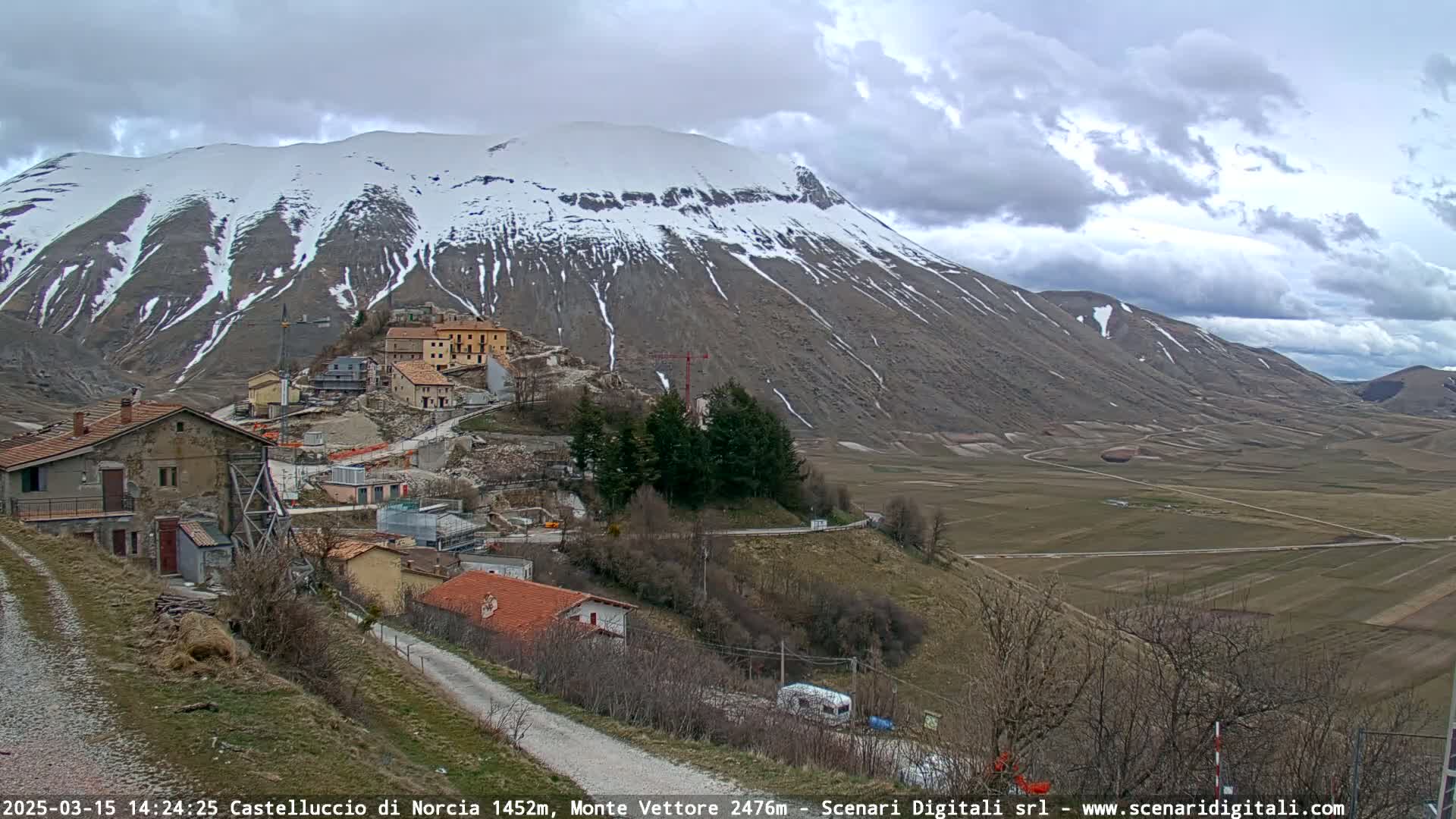 A snow-capped mountain overlooks a small village nestled in a valley under a cloudy sky.