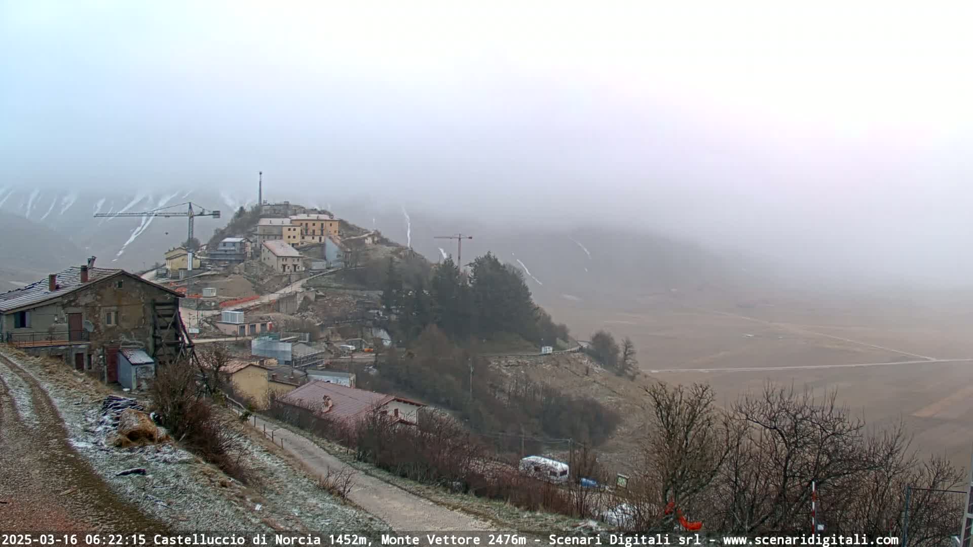 A foggy, snow-dusted hillside village with construction cranes and a flat, expansive valley in the background.