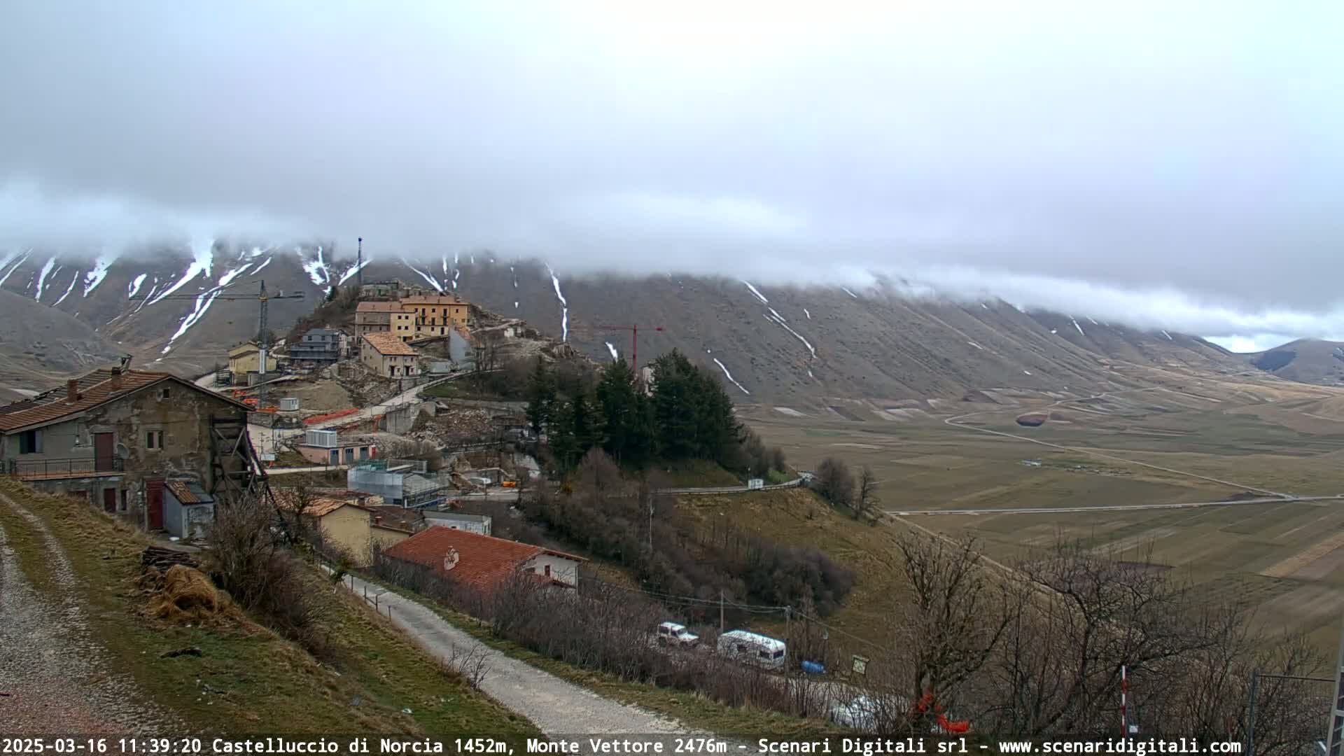 A small village nestled in a valley, partially obscured by fog, with snow-dusted mountains in the background.