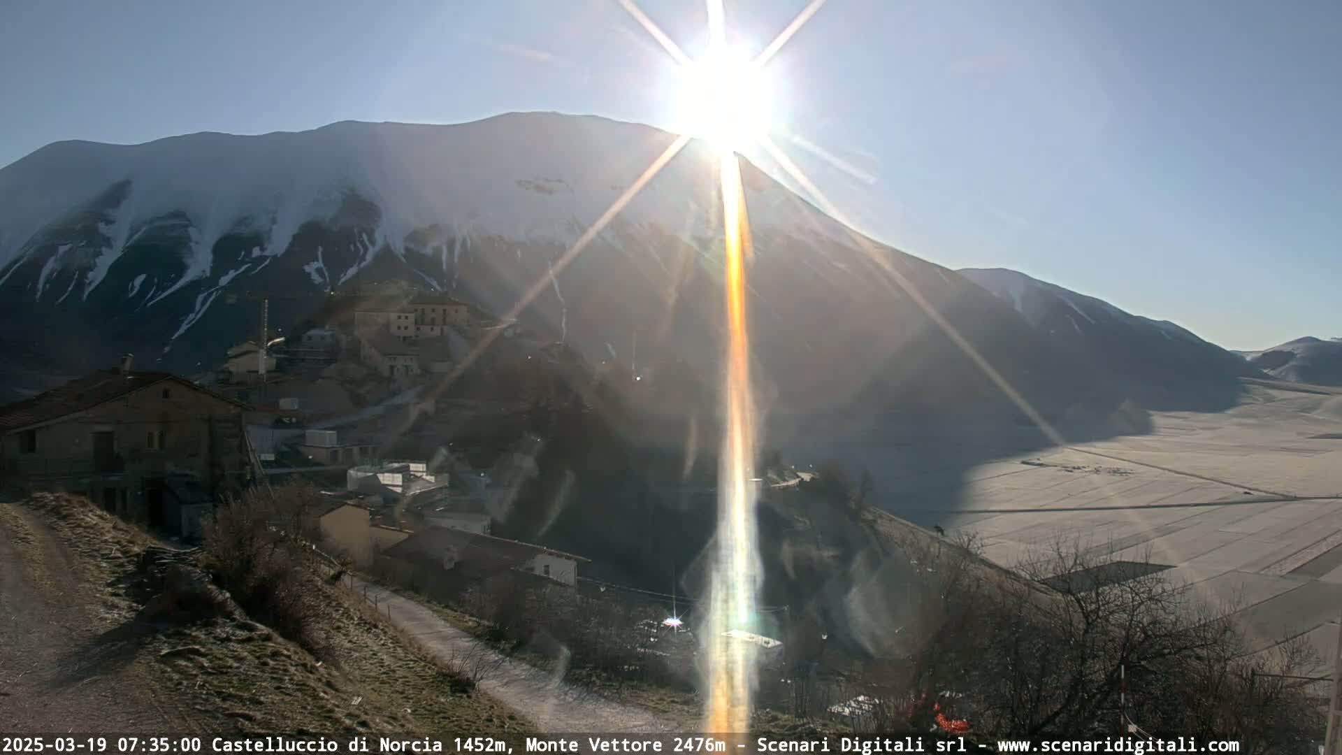 A sun-drenched mountainside village is nestled below a snow-capped mountain on a bright, clear day.