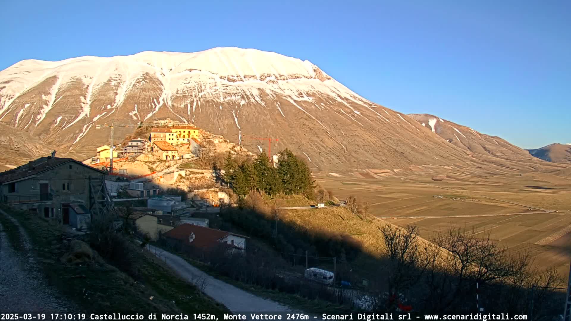 A snow-capped mountain overlooks a village nestled in a valley under a clear blue sky.