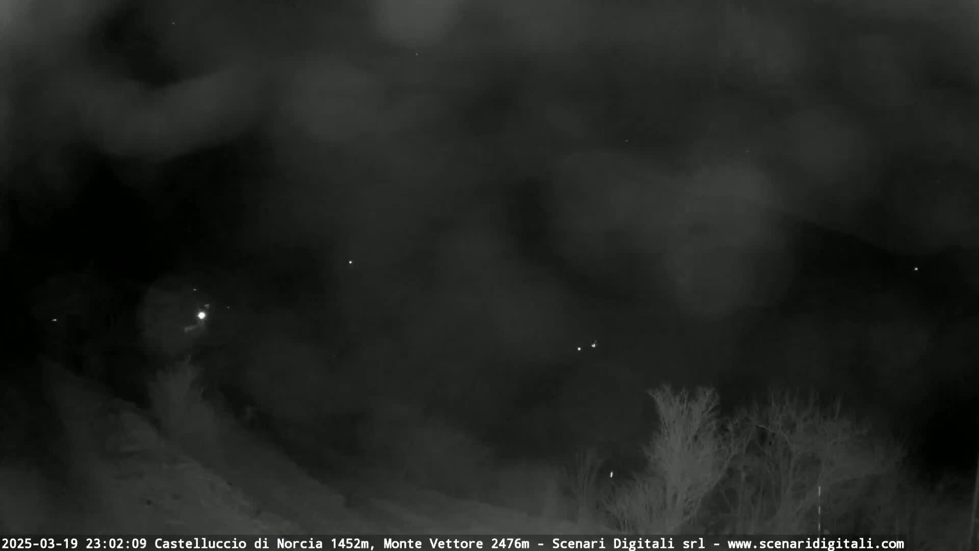 A nighttime, foggy landscape shows dark trees and hills under a cloudy sky, with a few distant lights visible.