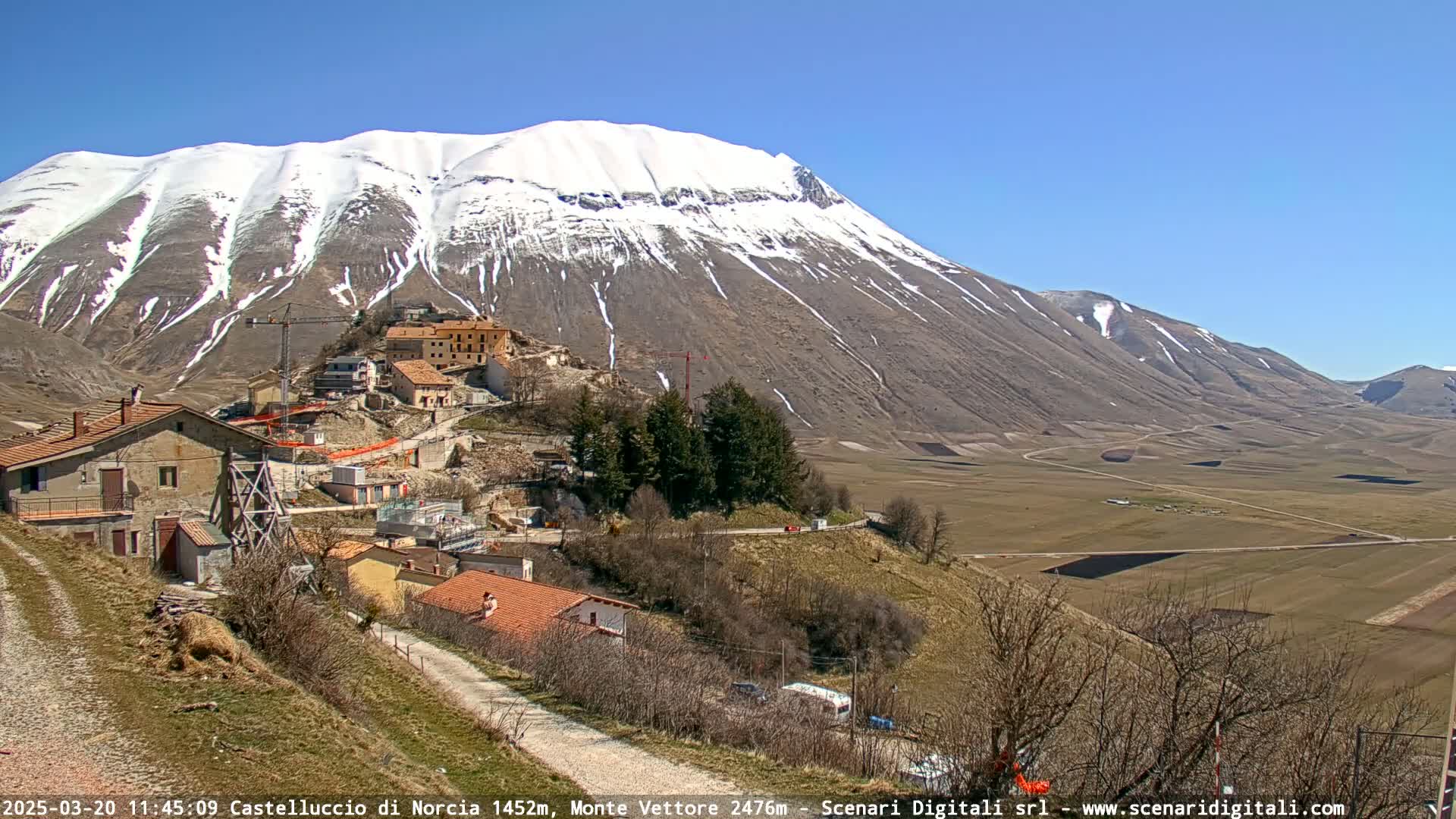 A snow-capped mountain overlooks a small village with ongoing construction, situated in a valley under a clear blue sky.