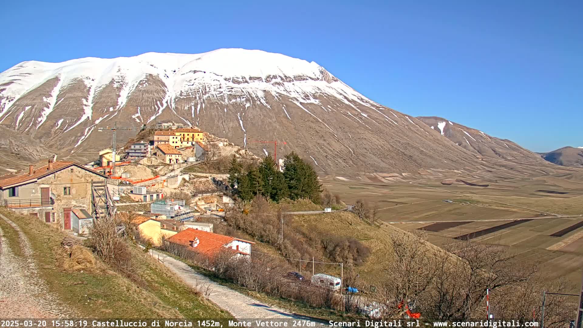 A small village nestled in a valley below a snow-capped mountain, under a clear blue sky.