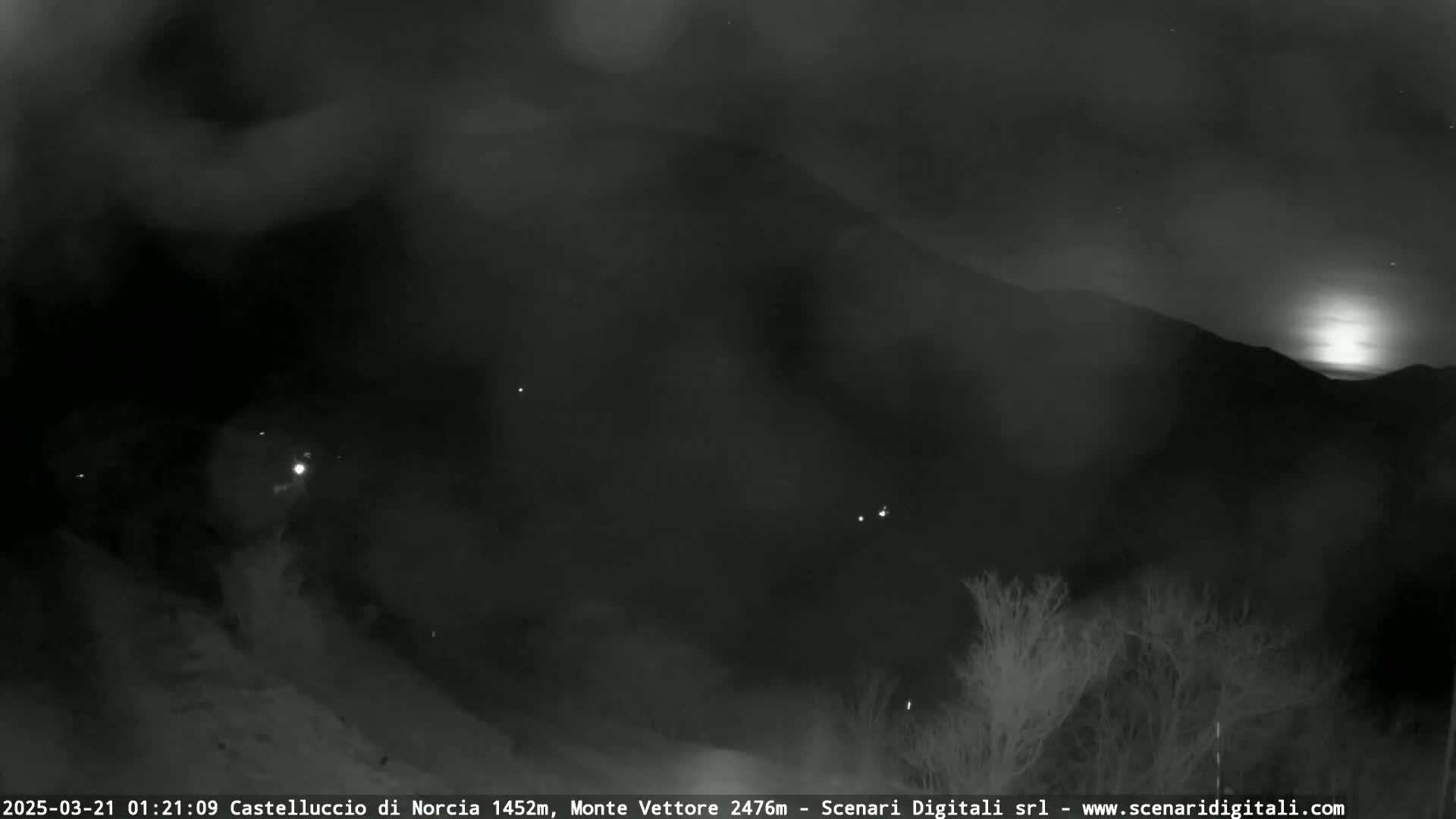 A nighttime, hazy view shows a moonlit mountain range with sparse vegetation and a few distant lights in the foreground.