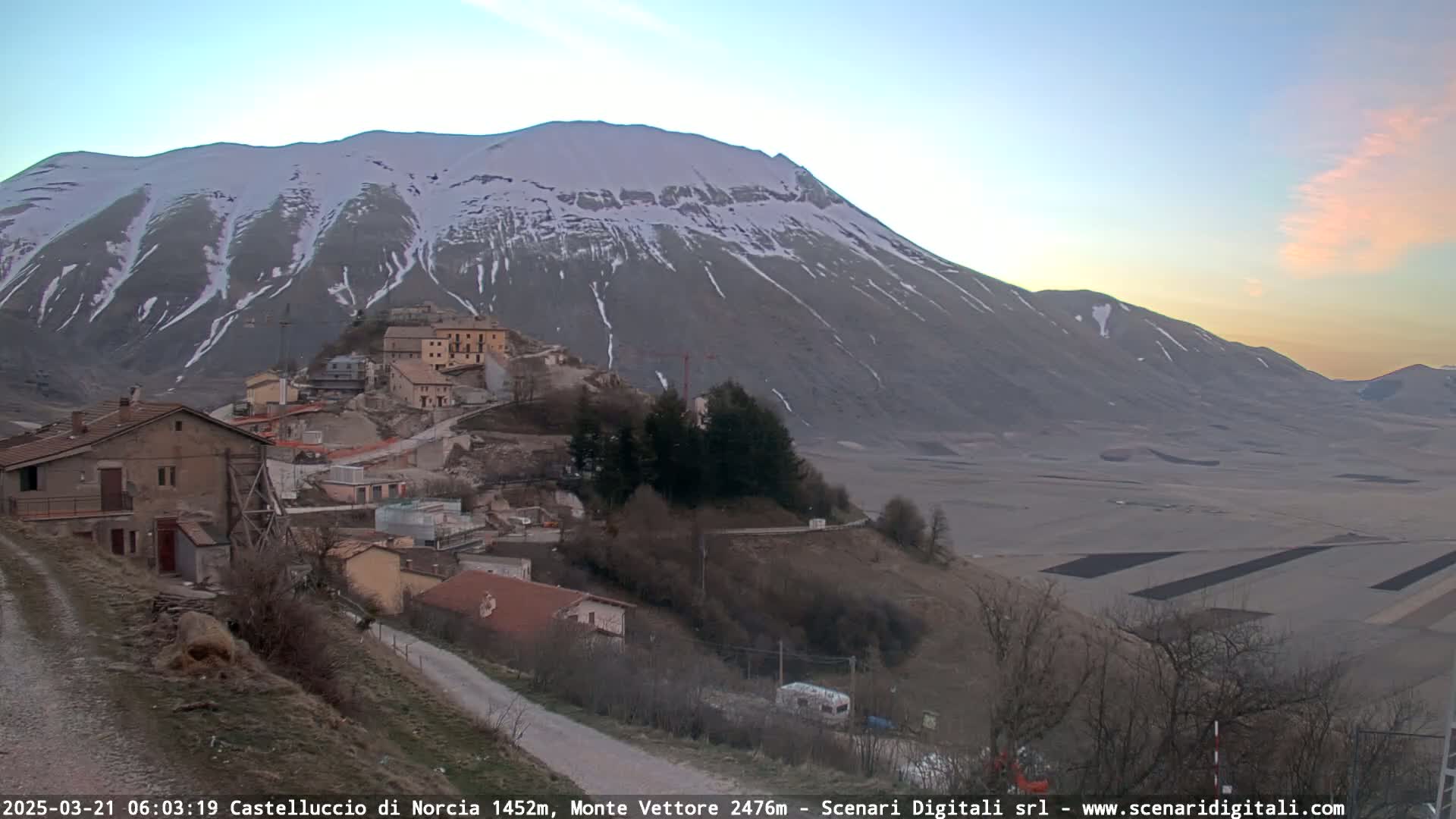A small village nestled on a hillside below a snow-capped mountain range at sunrise, under a mostly clear sky.