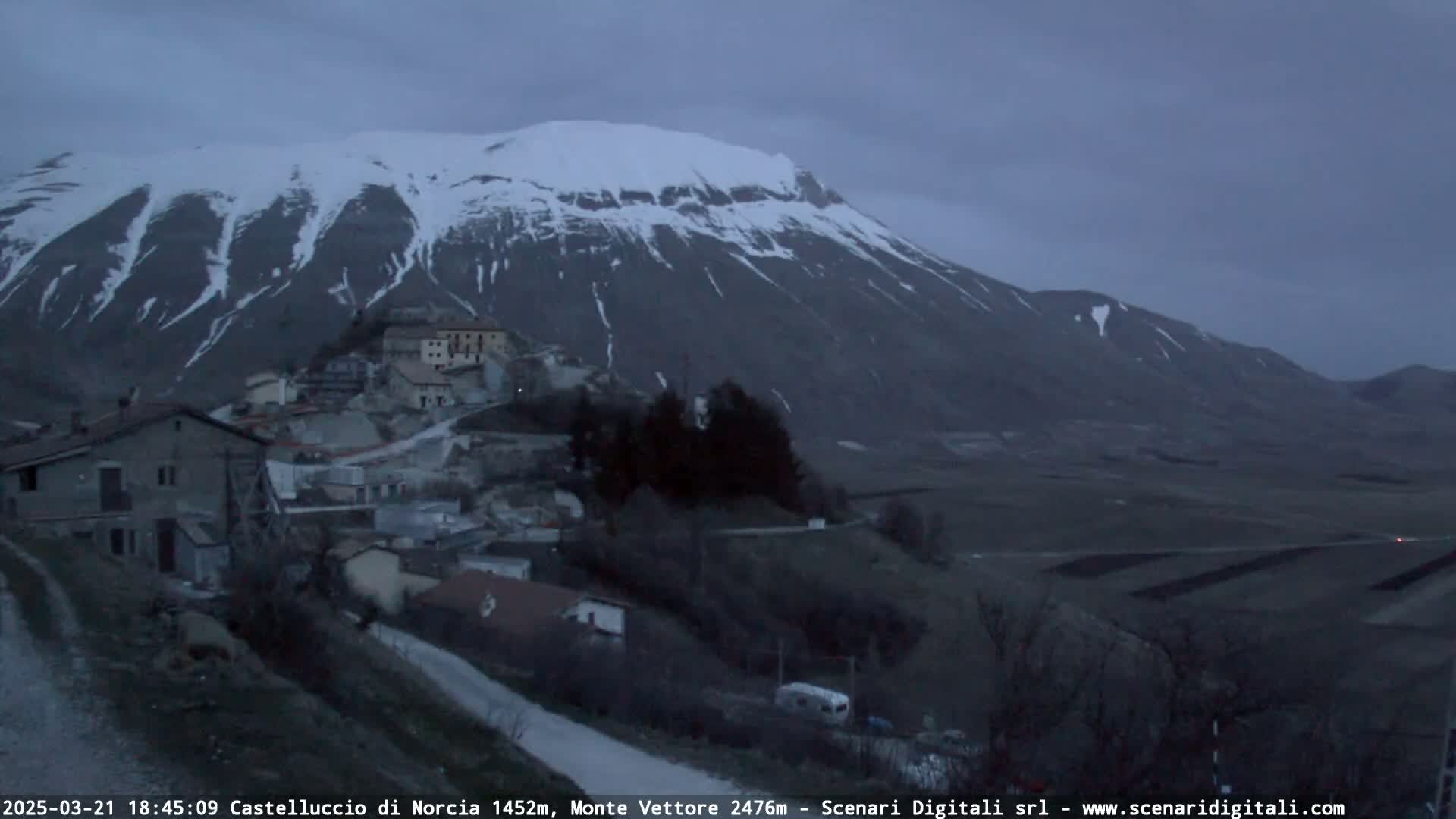 A snow-capped mountain looms over a small village nestled in a valley under a dusky sky.