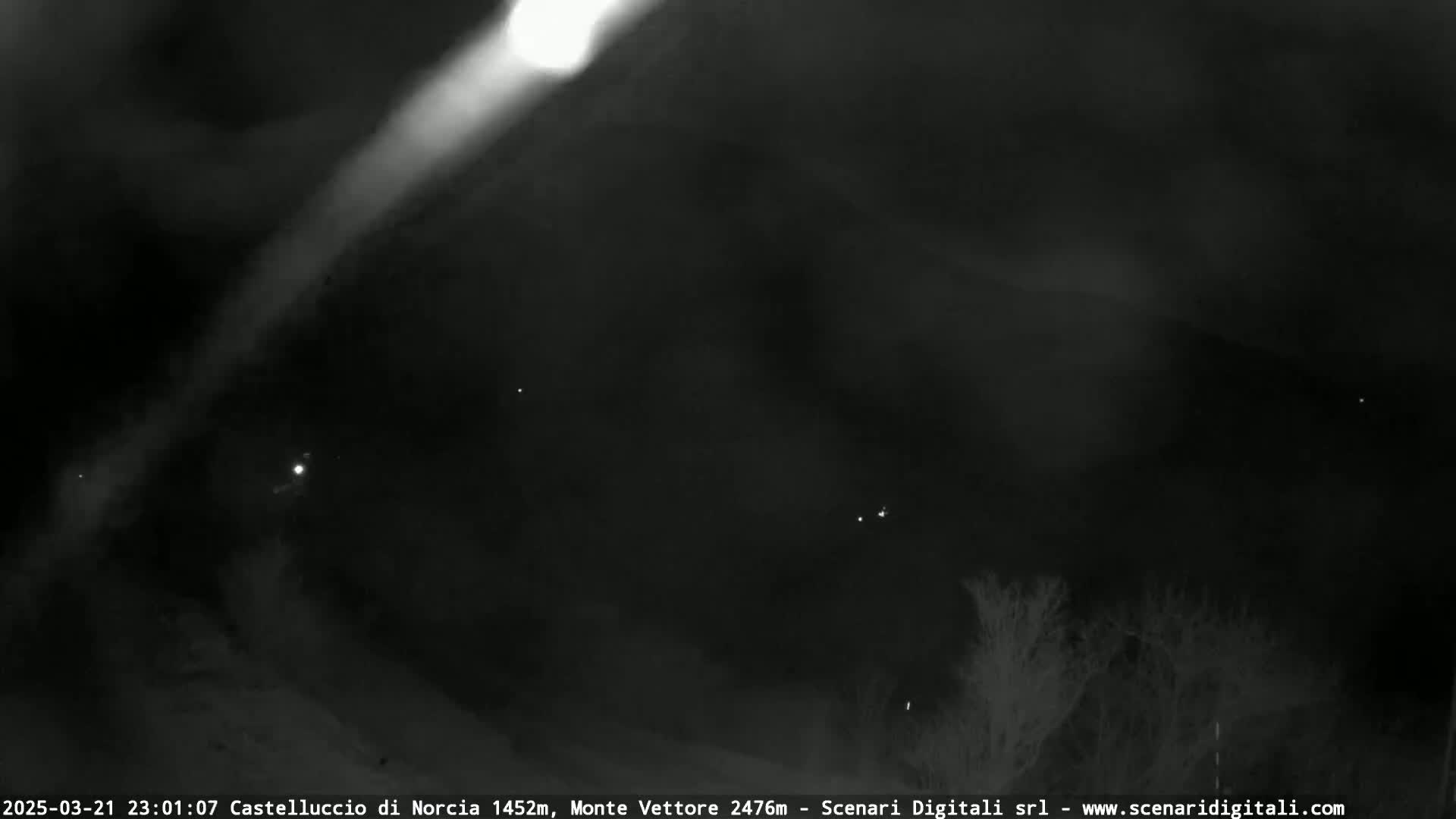 A nighttime, low-light outdoor image shows a bright beam of light angled across a dark, mostly cloudy sky above a dark landscape with a few faint lights.
