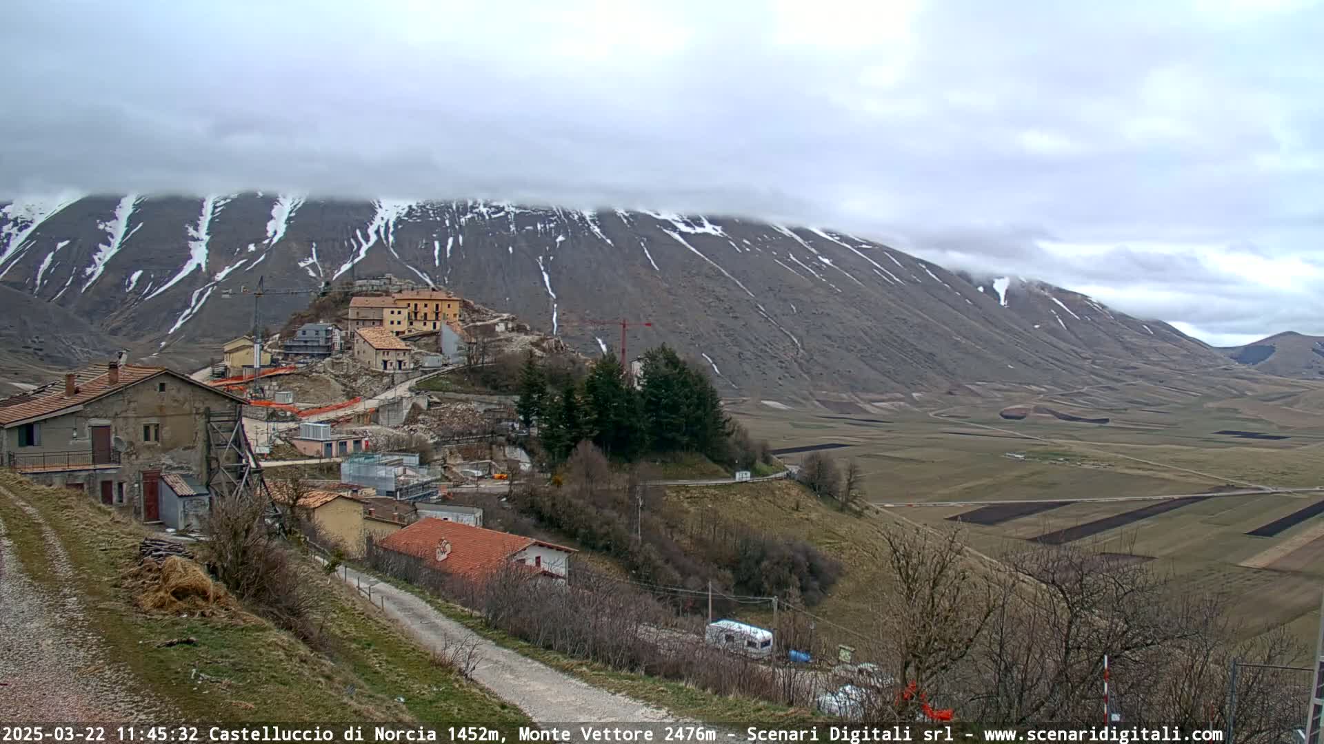 A small village nestled on a hillside overlooks a vast, flat plain, with a snow-capped mountain in the background under a cloudy sky.