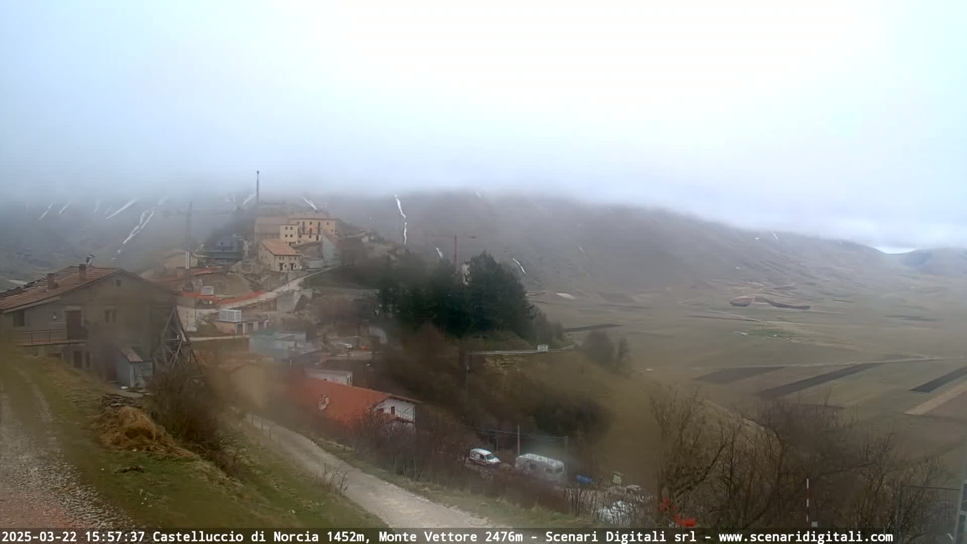 A foggy, overcast day reveals a small village nestled in a valley, with distant mountains partially obscured by mist and patches of snow visible on higher elevations.