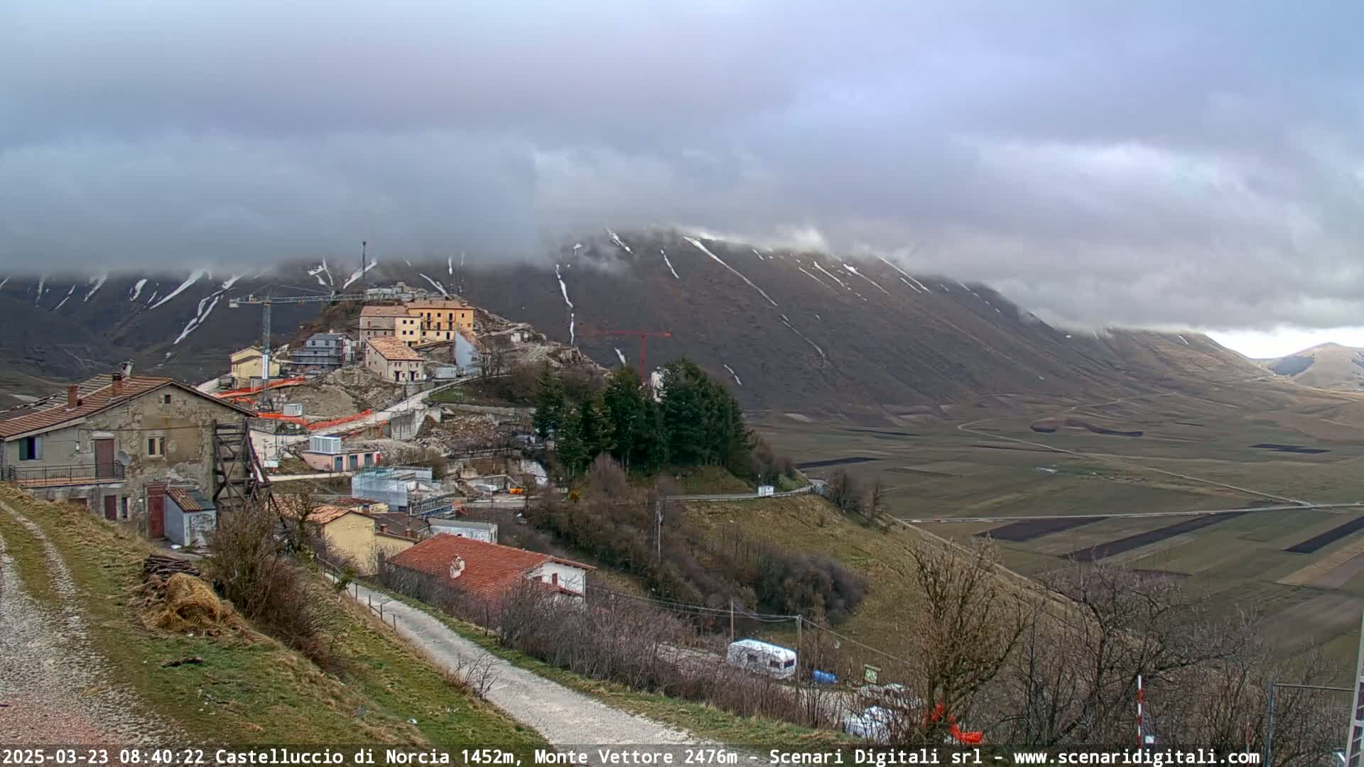 A small village nestled in a valley, with a partially cloudy sky and snow-dusted mountains in the background.