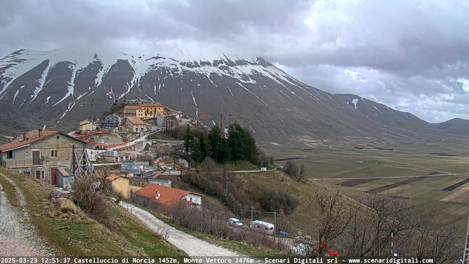 A small village nestled on a hillside beneath a snow-capped mountain, on a partly cloudy day, overlooks a vast, flat valley of farmland.
