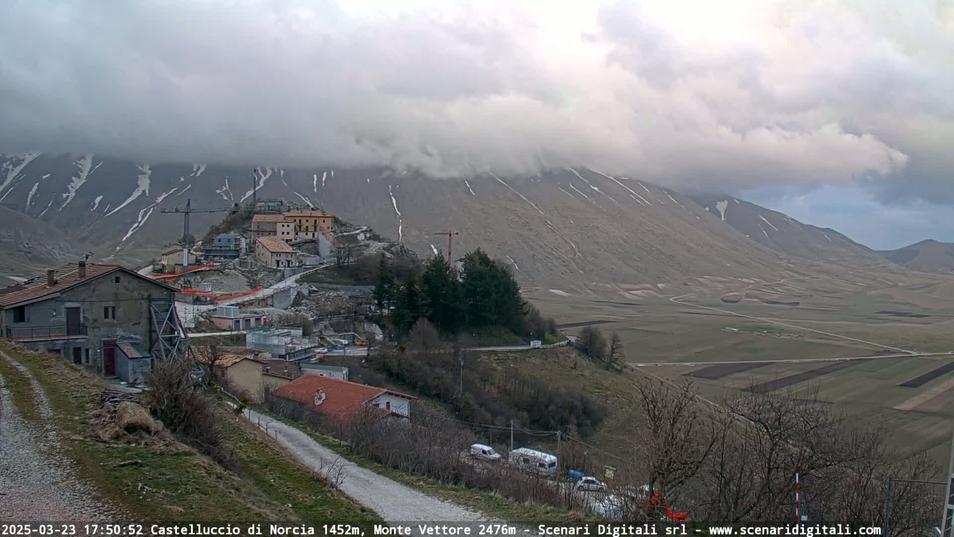 A partly cloudy day reveals a mountain village with construction underway, nestled in a valley with snow-capped mountains in the background and farmland stretching into the distance.