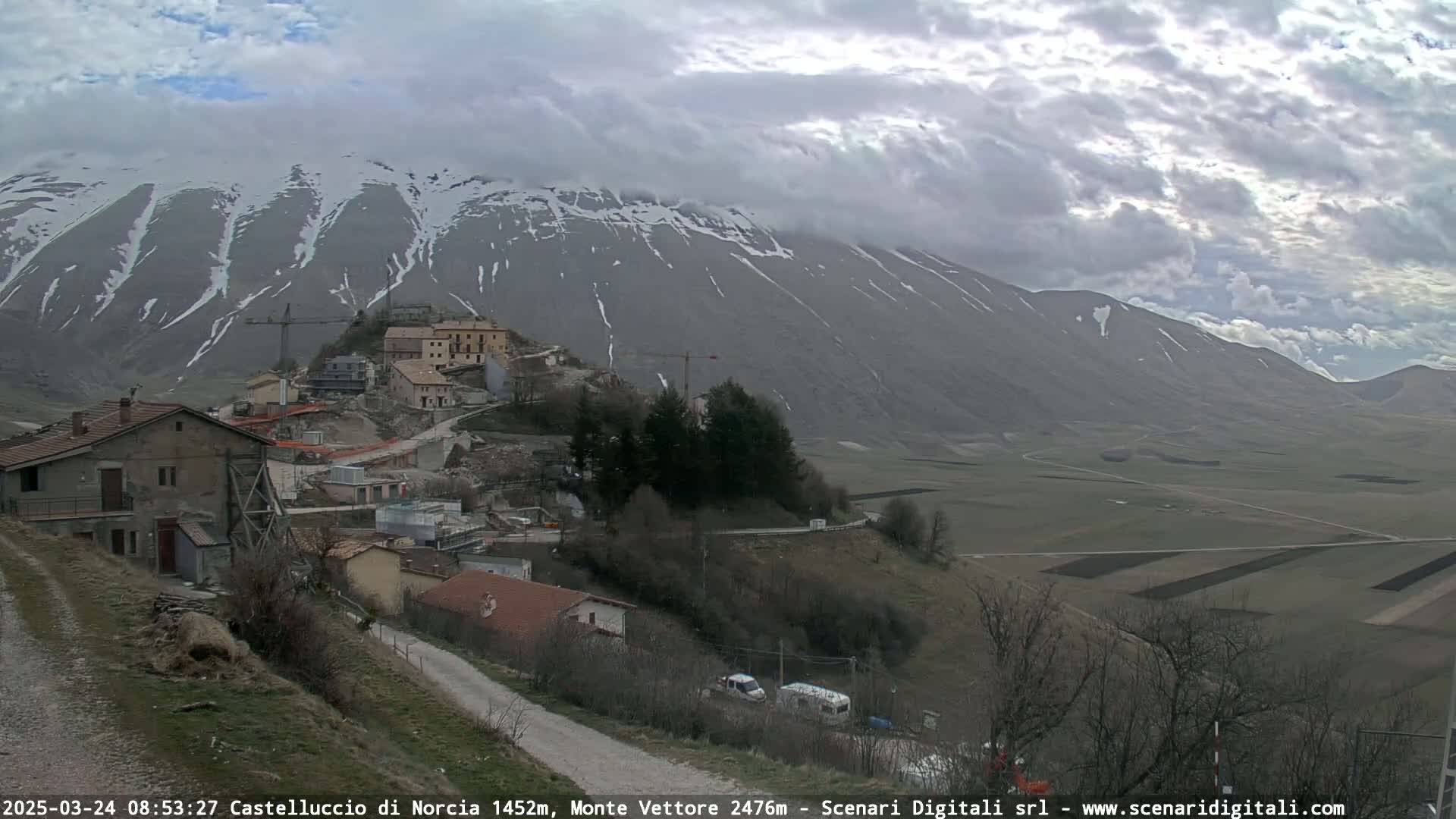 A partially cloudy day reveals a snow-dusted mountain overlooking a small village nestled in a valley of farmland.