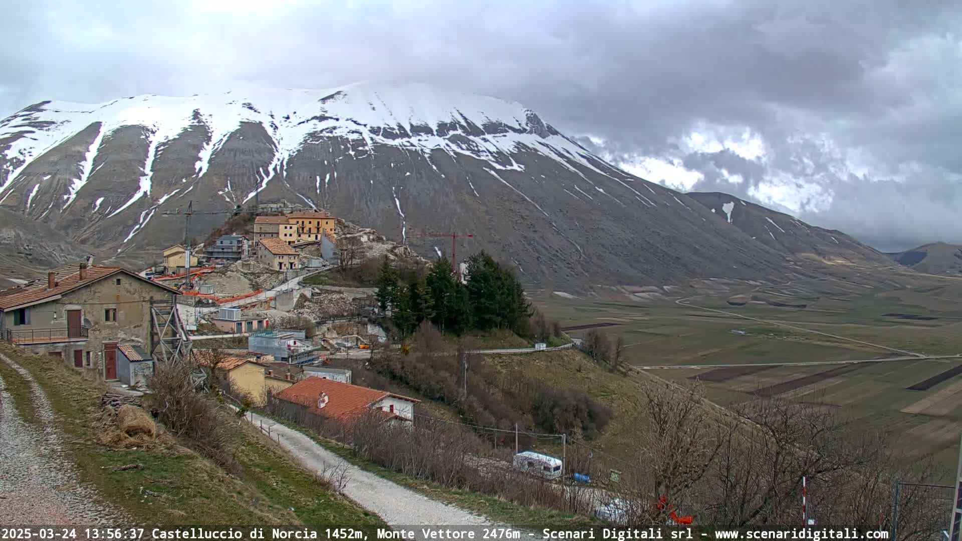 A snow-capped mountain overlooks a small village nestled in a valley under a cloudy sky.