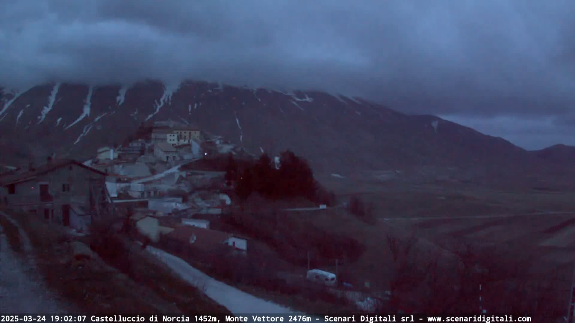 A small village nestled in a valley below a large, snow-dusted mountain under a cloudy, dark sky.