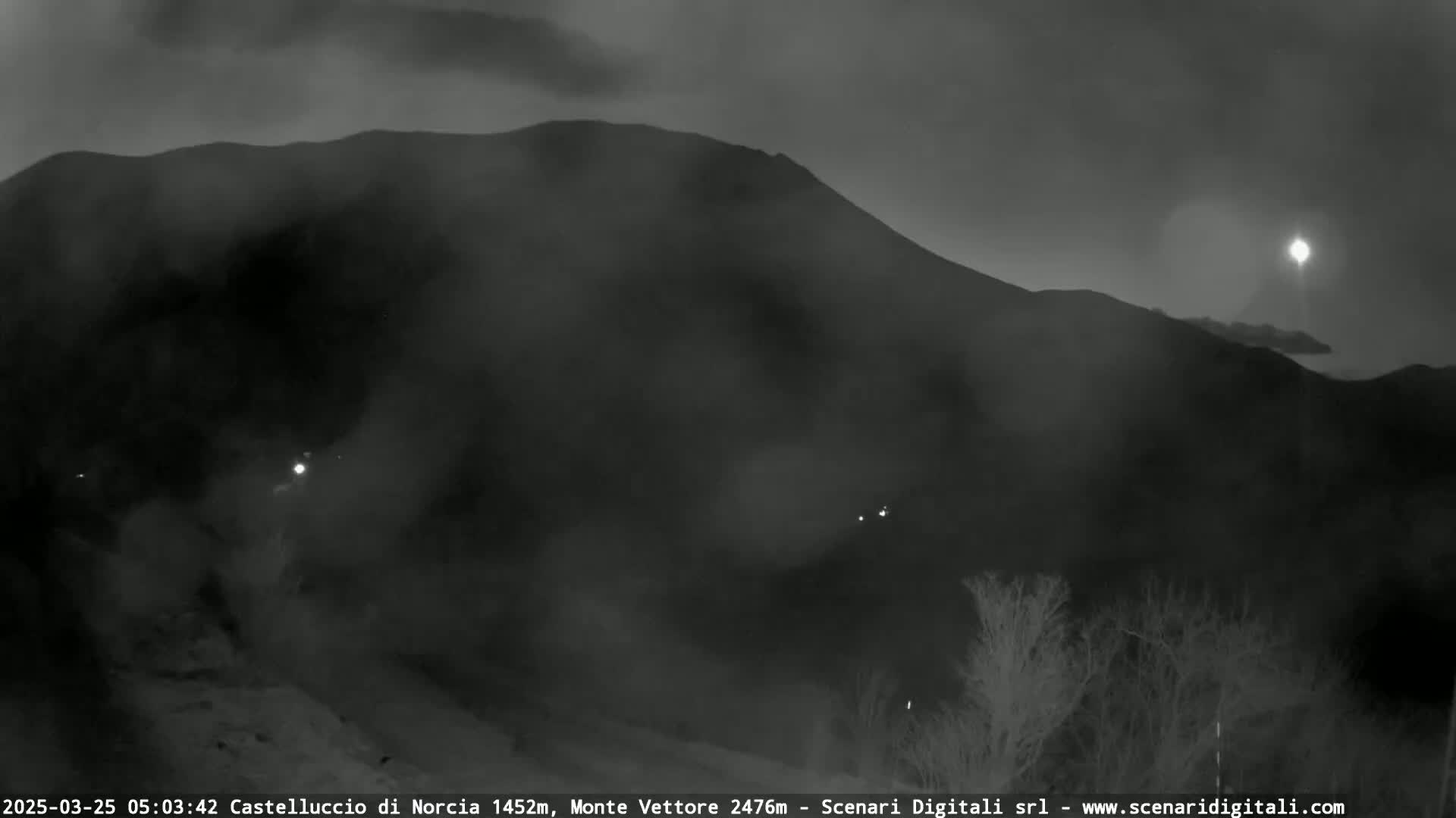 A dark, foggy mountain range is visible at night, with a few faint lights scattered in the foreground and a bright light in the far distance.