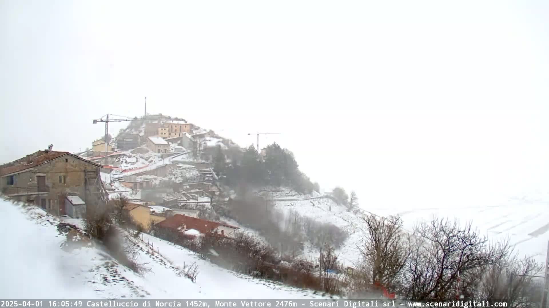 A snow-covered hillside village with construction cranes visible, under a heavy snowfall.