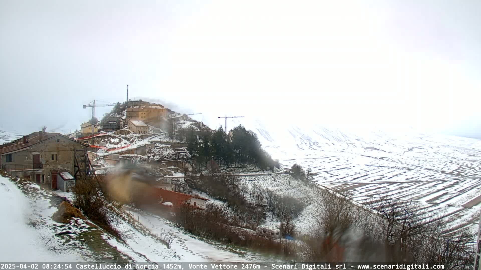 A snow-covered village with construction cranes near a hilltop fortress is visible under an overcast sky.