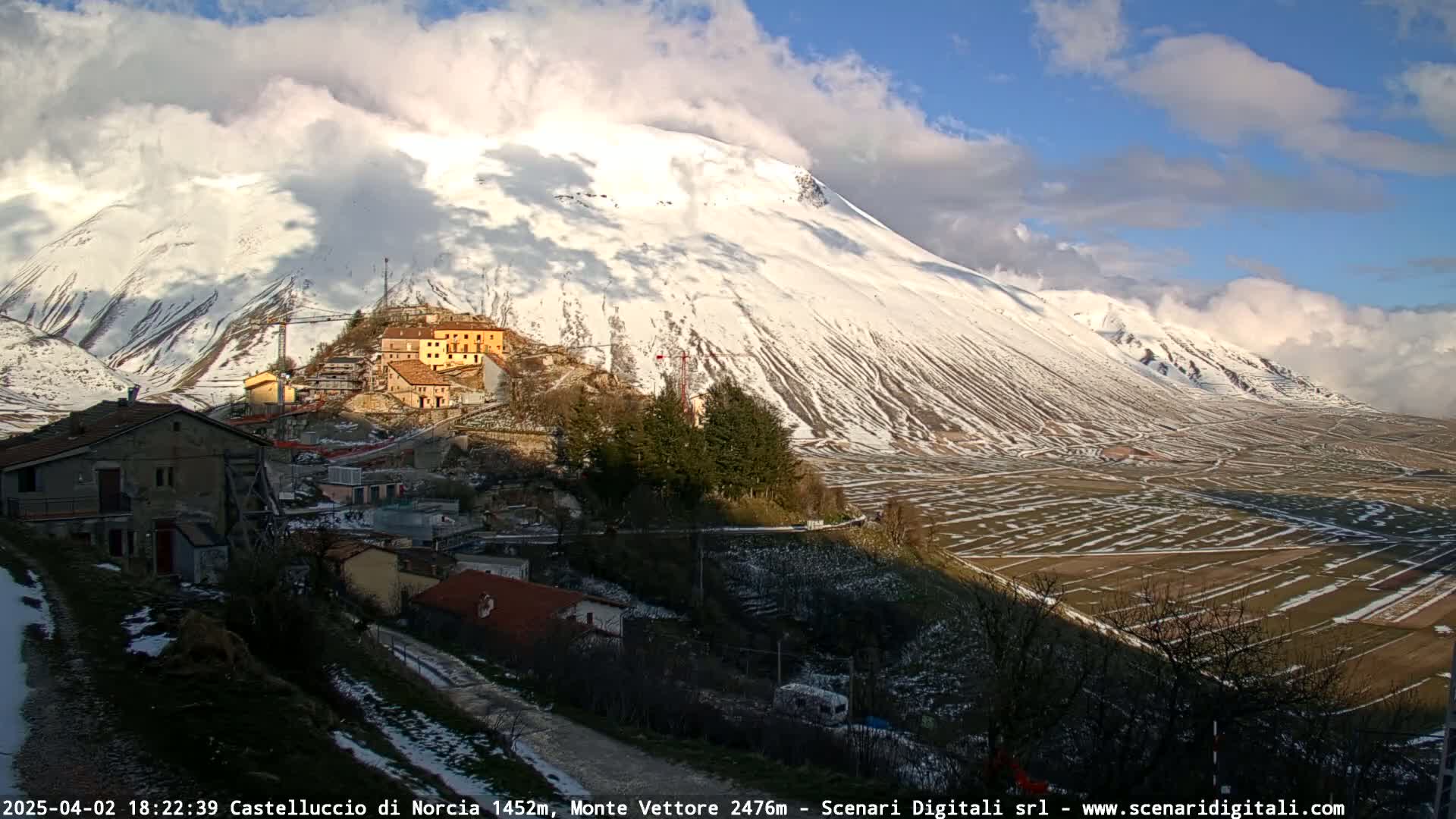 A snow-covered mountain looms over a small village nestled in a valley under a partly cloudy sky.