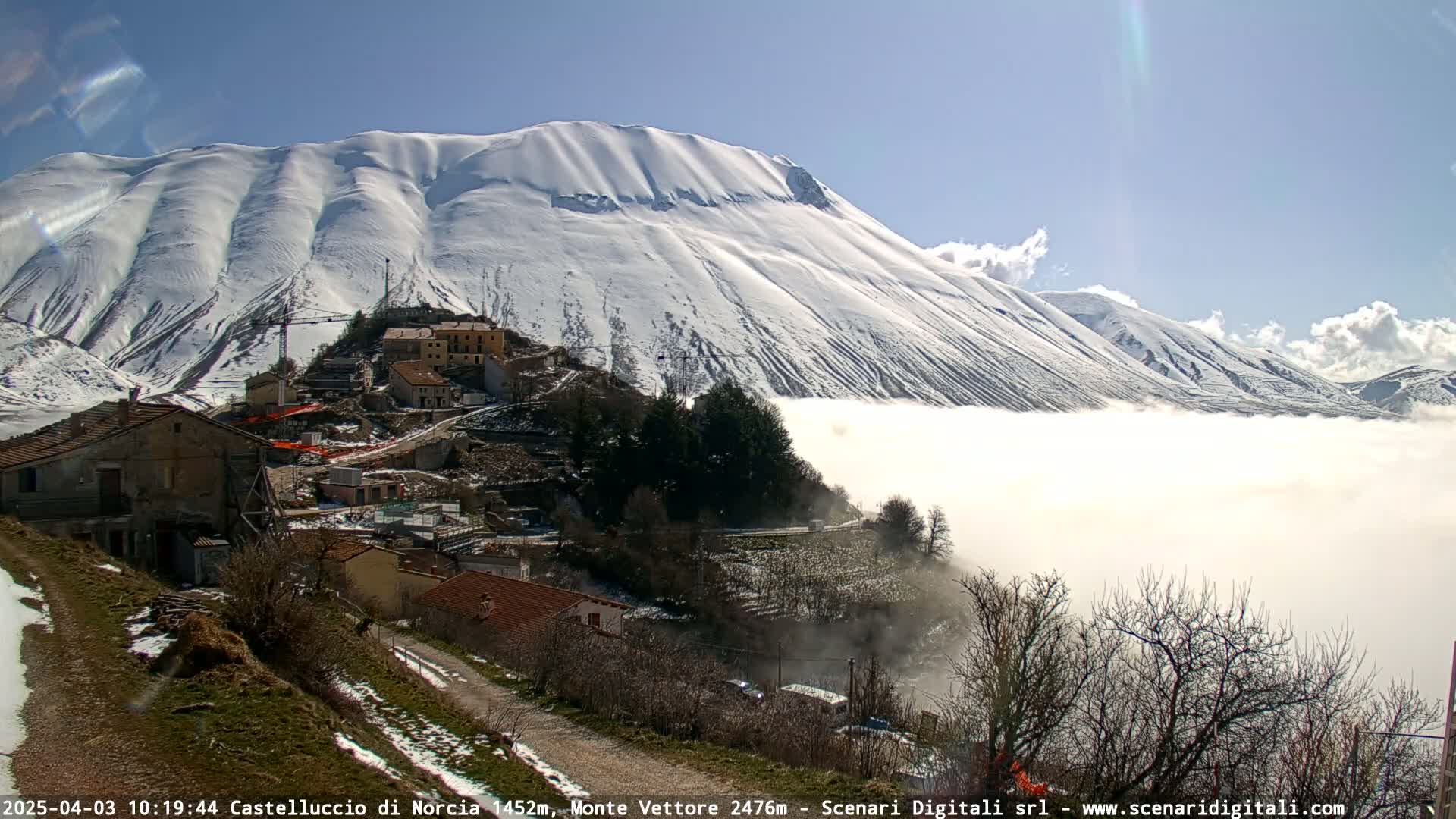 A snow-covered mountain looms over a village nestled in a valley shrouded in fog under a mostly sunny sky.