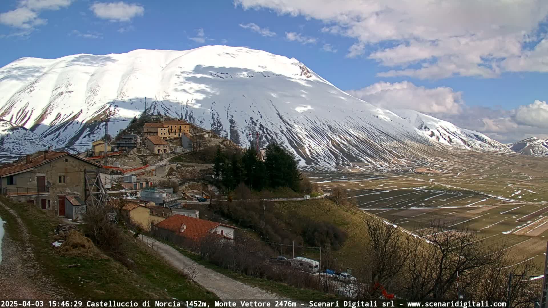 A small village nestled on a hillside overlooks a valley of farmland and a large snow-covered mountain under a partly cloudy sky.