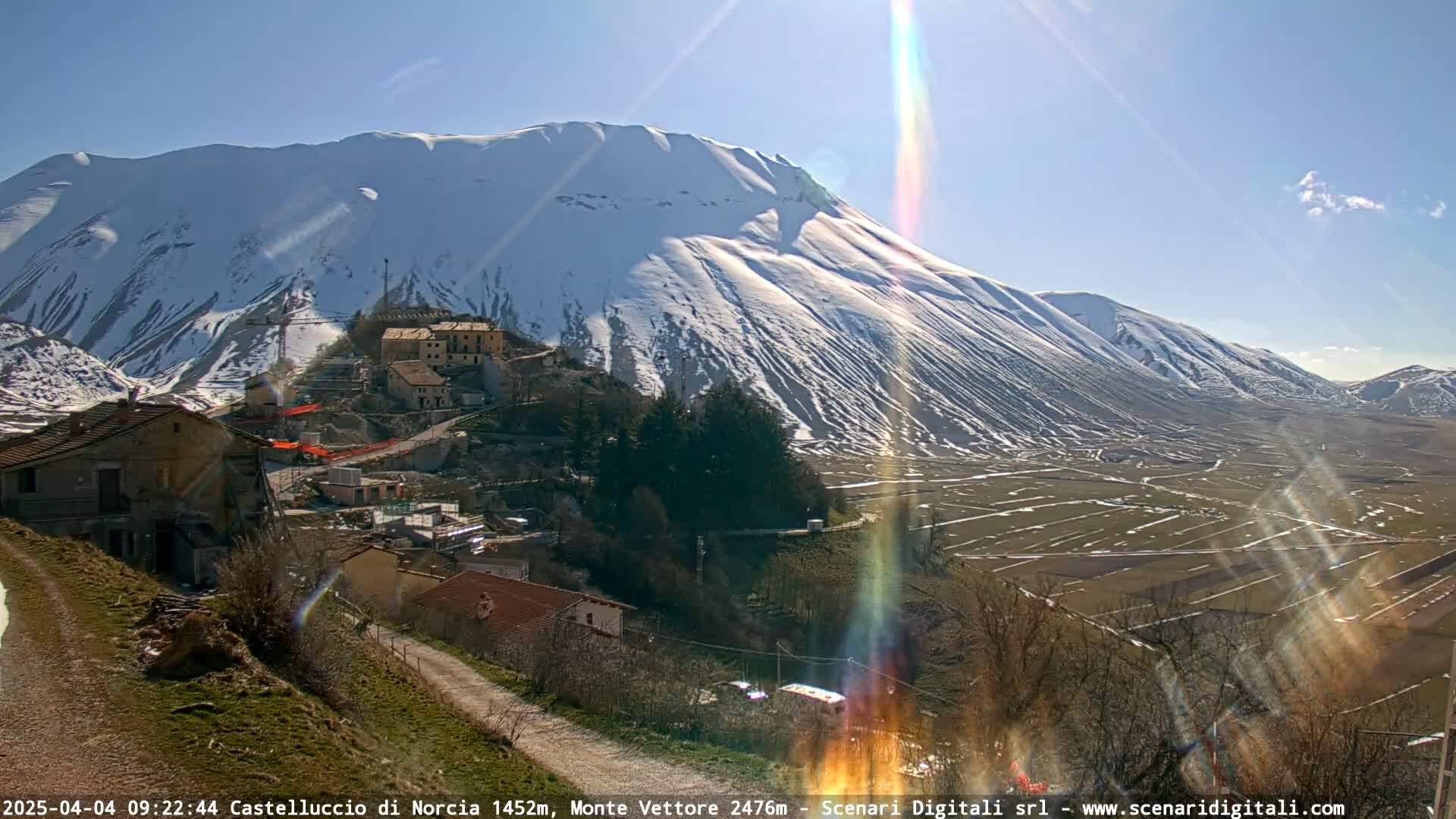 A sunny day reveals a snow-capped mountain overlooking a valley of farmland and a small village, with sunbeams streaking across the landscape.