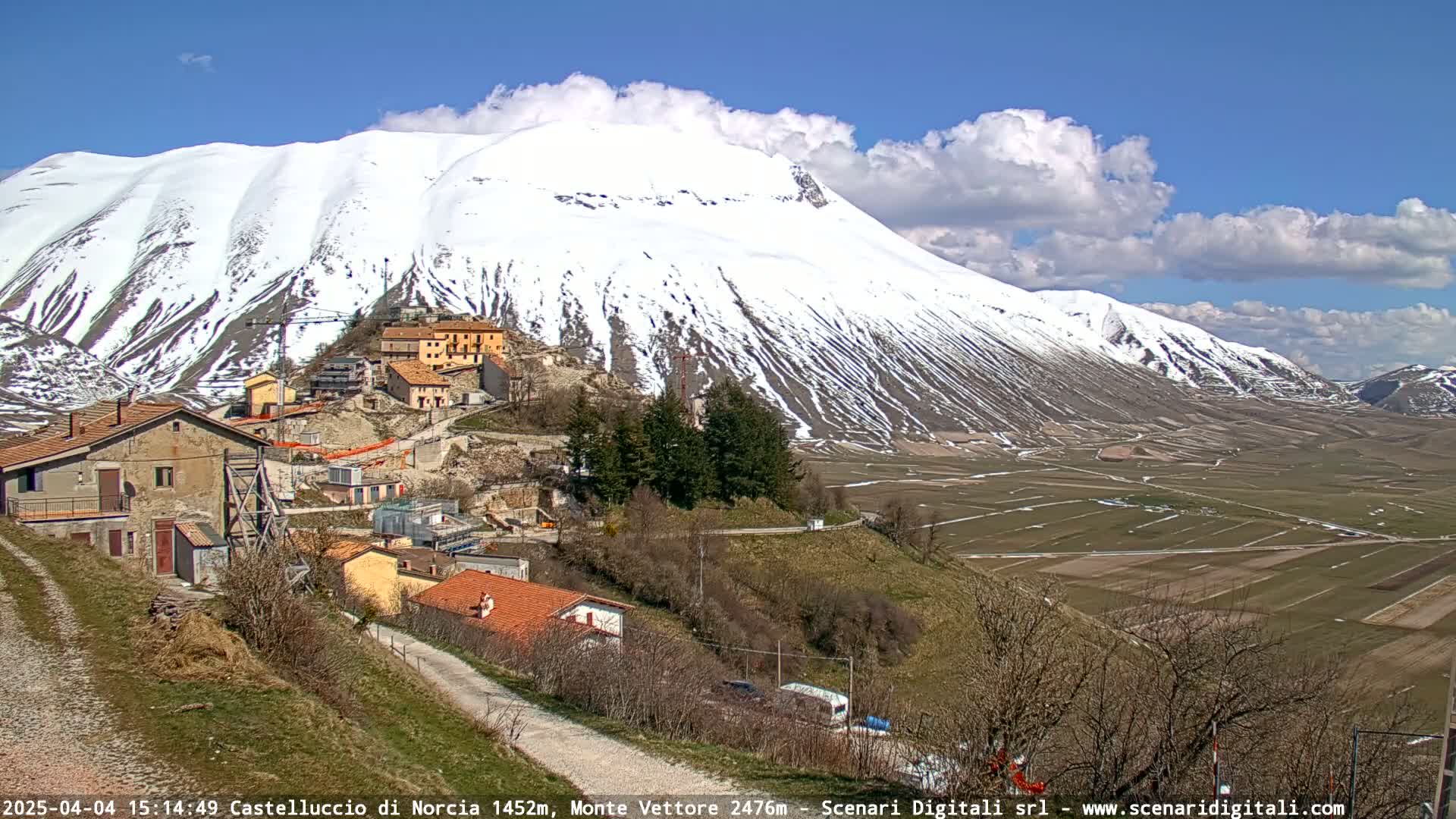 A small village nestled in a valley, with a large snow-capped mountain in the background under a partly cloudy sky.