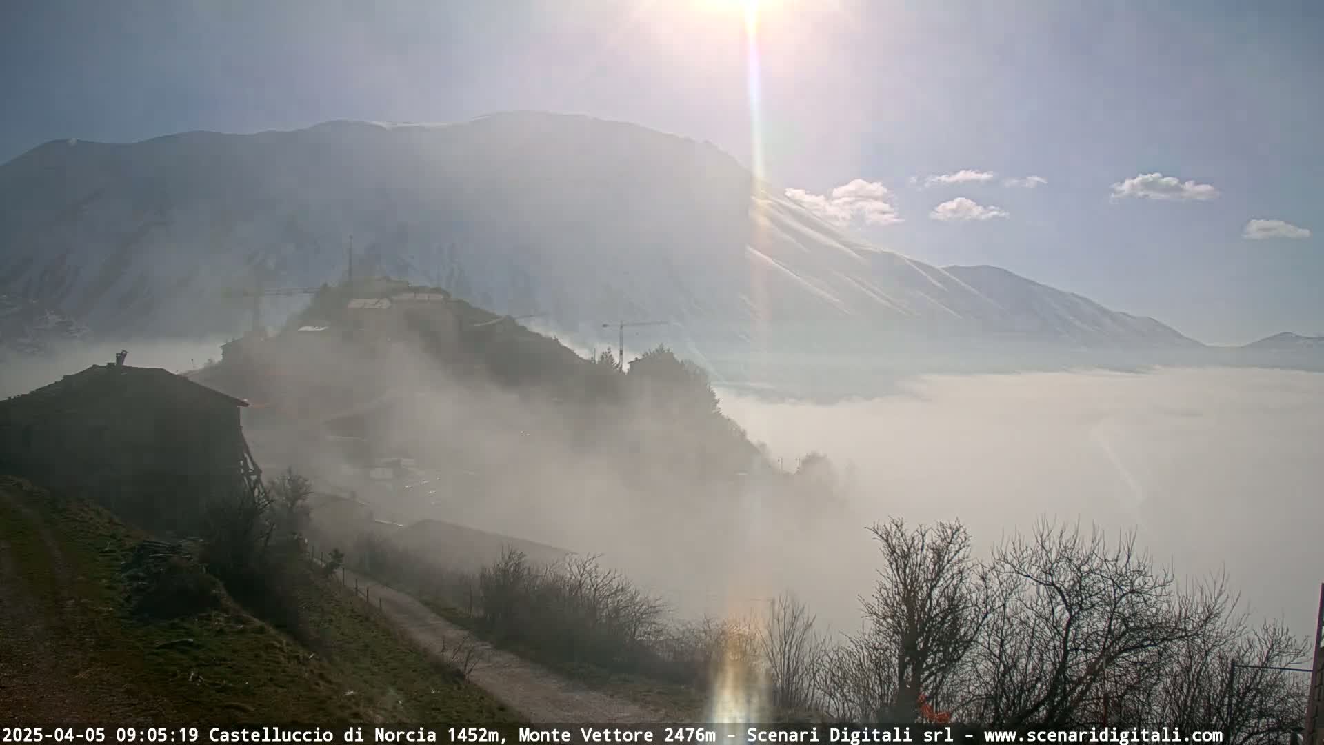 A foggy valley scene reveals a village nestled in the hills, with snow-capped mountains in the background under a bright, sunny sky.