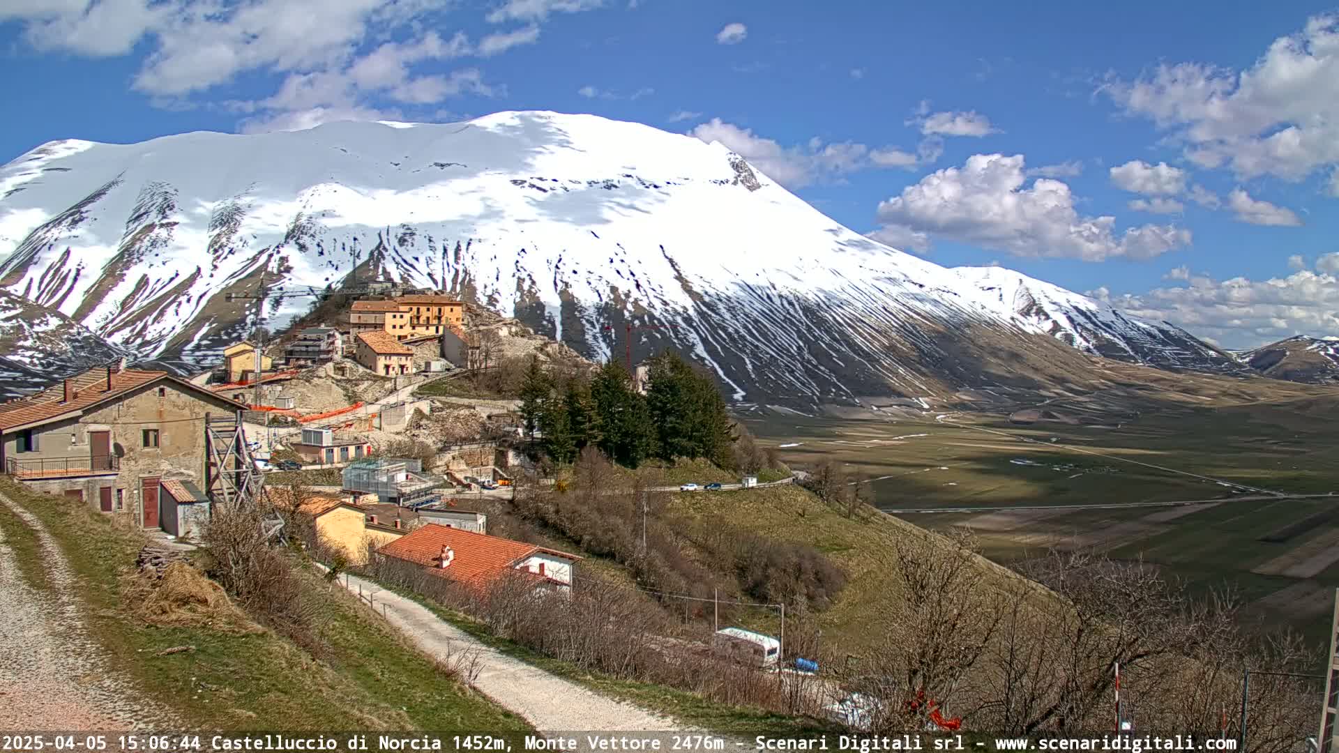 A small village nestled on a hillside overlooks a valley and a large, snow-capped mountain under a partly cloudy sky.