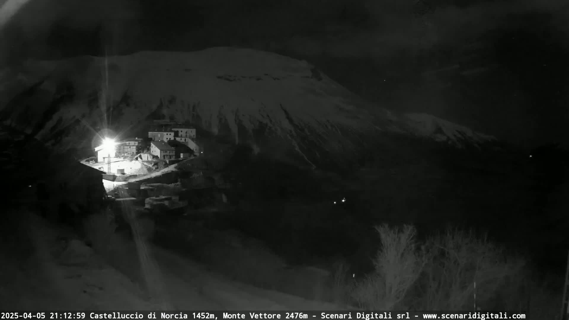A nighttime, snow-covered mountain looms over a small, illuminated village.