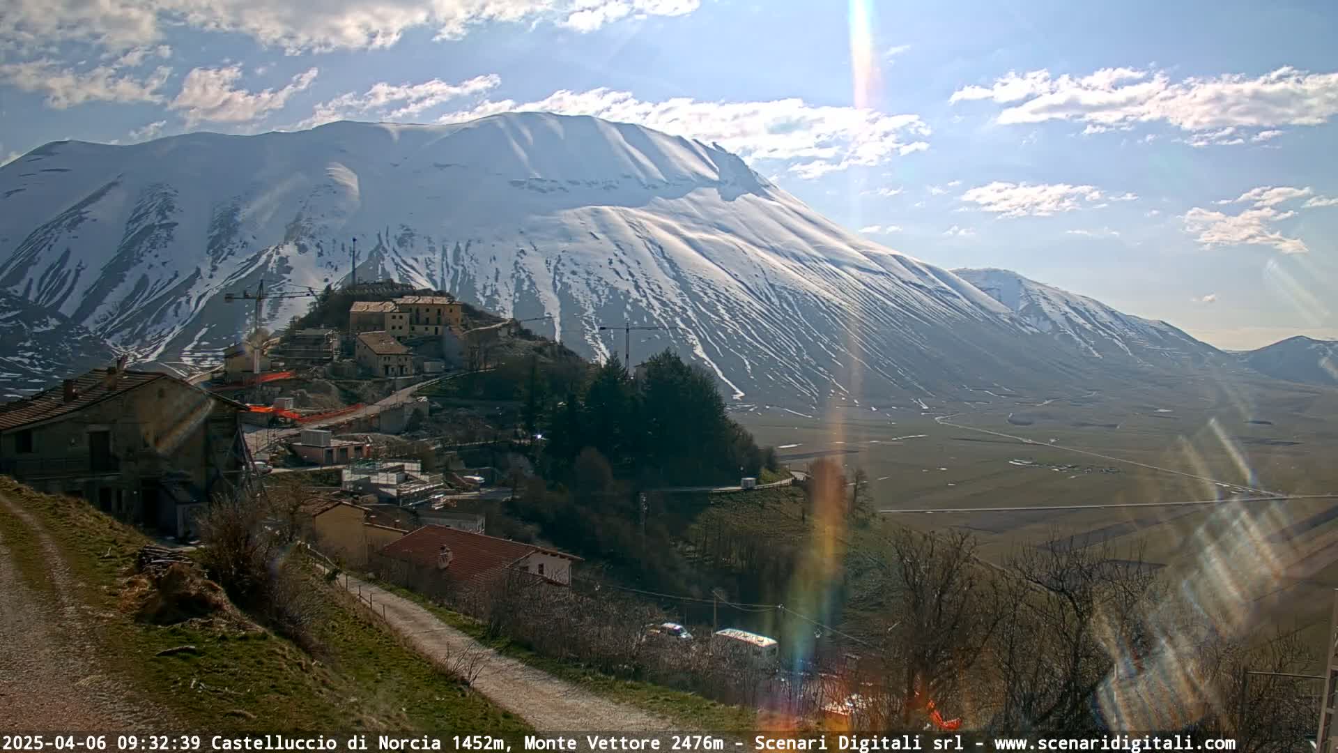 A partly sunny day reveals a snow-capped mountain behind a small village nestled in a valley, with construction equipment visible near buildings on a hillside.