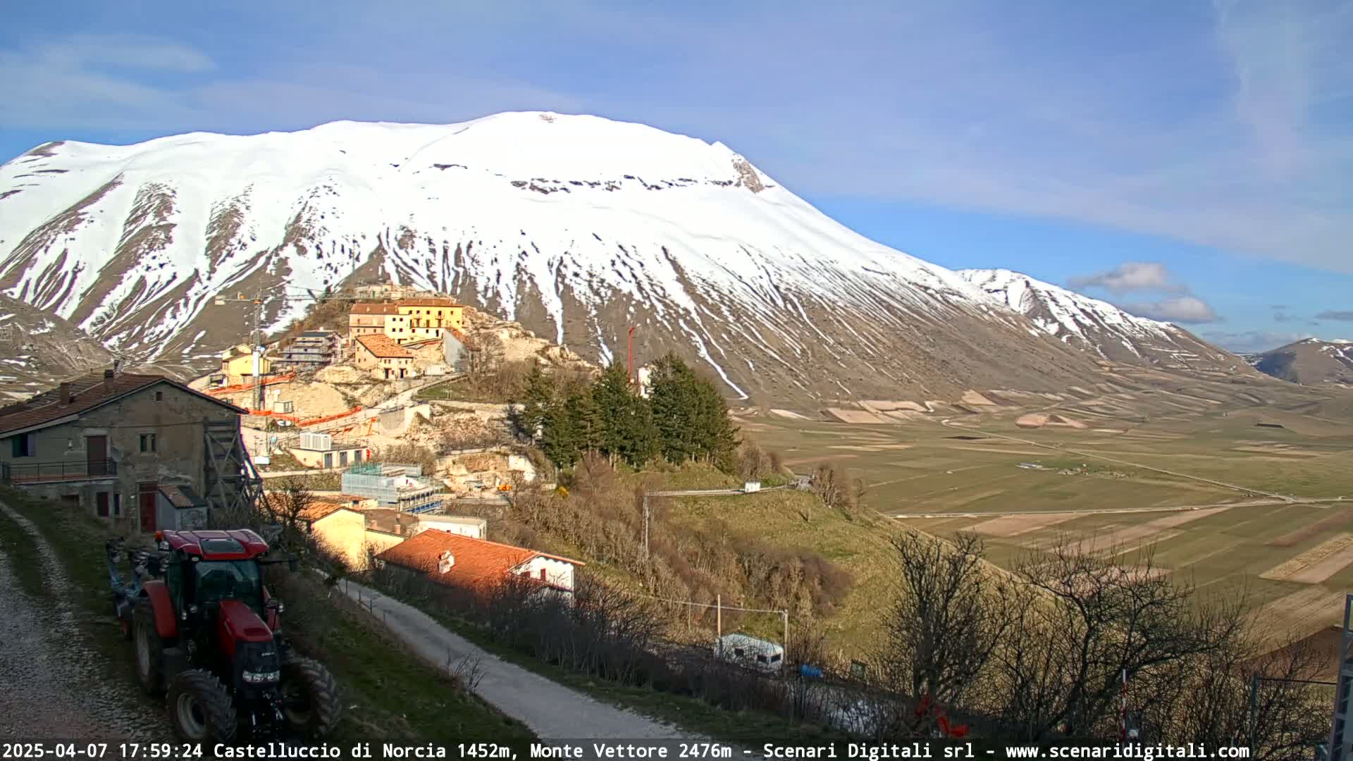 A tractor sits on a road overlooking a village nestled against a snow-capped mountain under a mostly sunny sky.