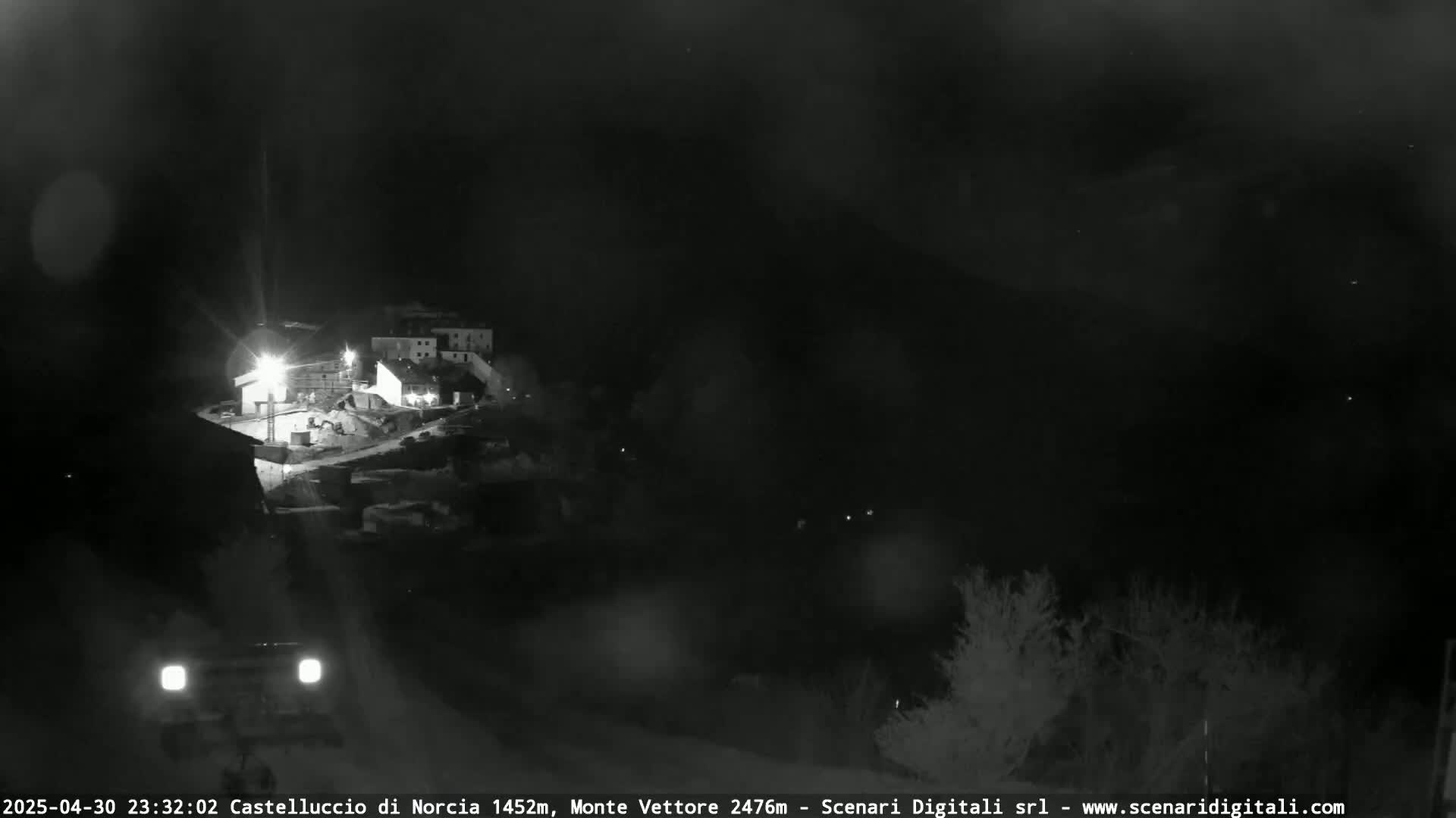 A nighttime, possibly rainy, view of a small village on a hillside, with a vehicle visible in the foreground.