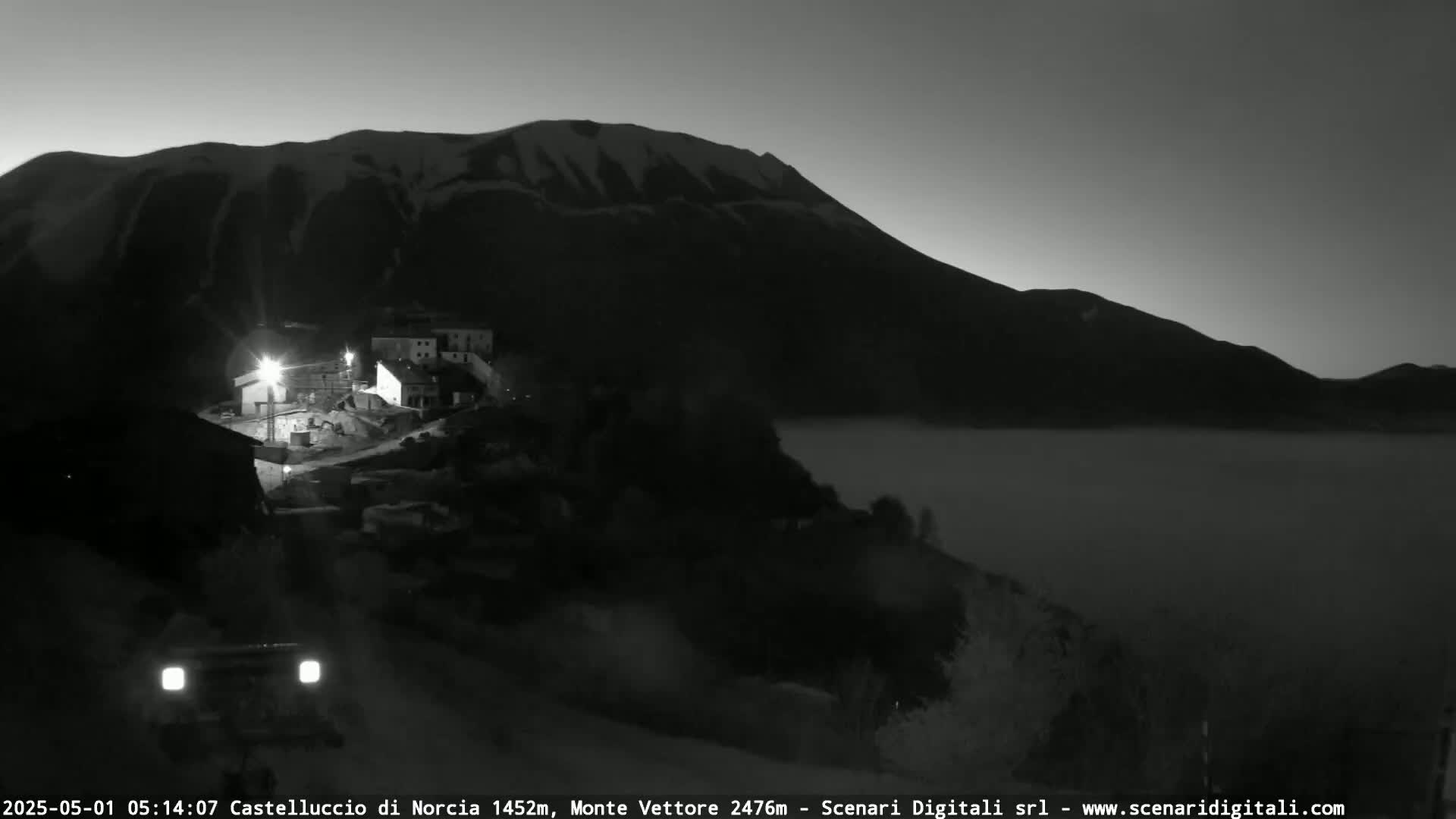 A nighttime, low-light view shows a small village nestled at the base of a snow-capped mountain, with fog or low clouds filling the valley below.