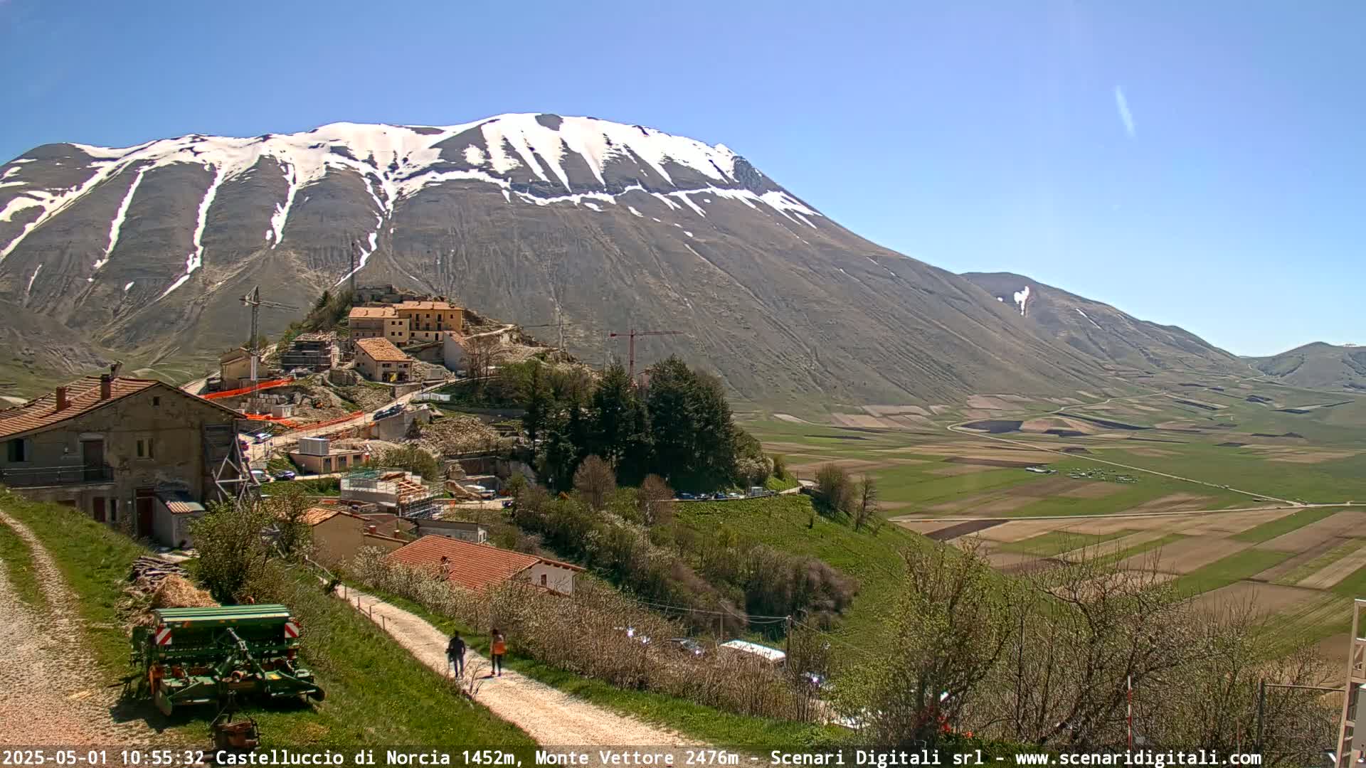 A partly snowy mountain overlooks a rural village and farmland under a clear blue sky.