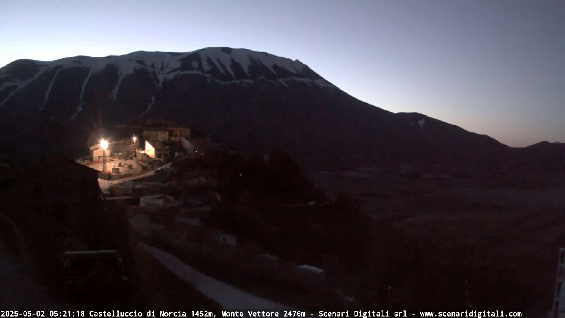 A small, dimly lit village sits at the base of a large, snow-capped mountain under a twilight sky.