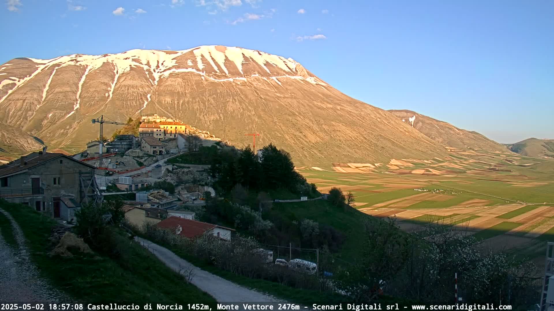 A partly snow-covered mountain overlooks a valley of farmland and a small town under a clear, sunny sky.