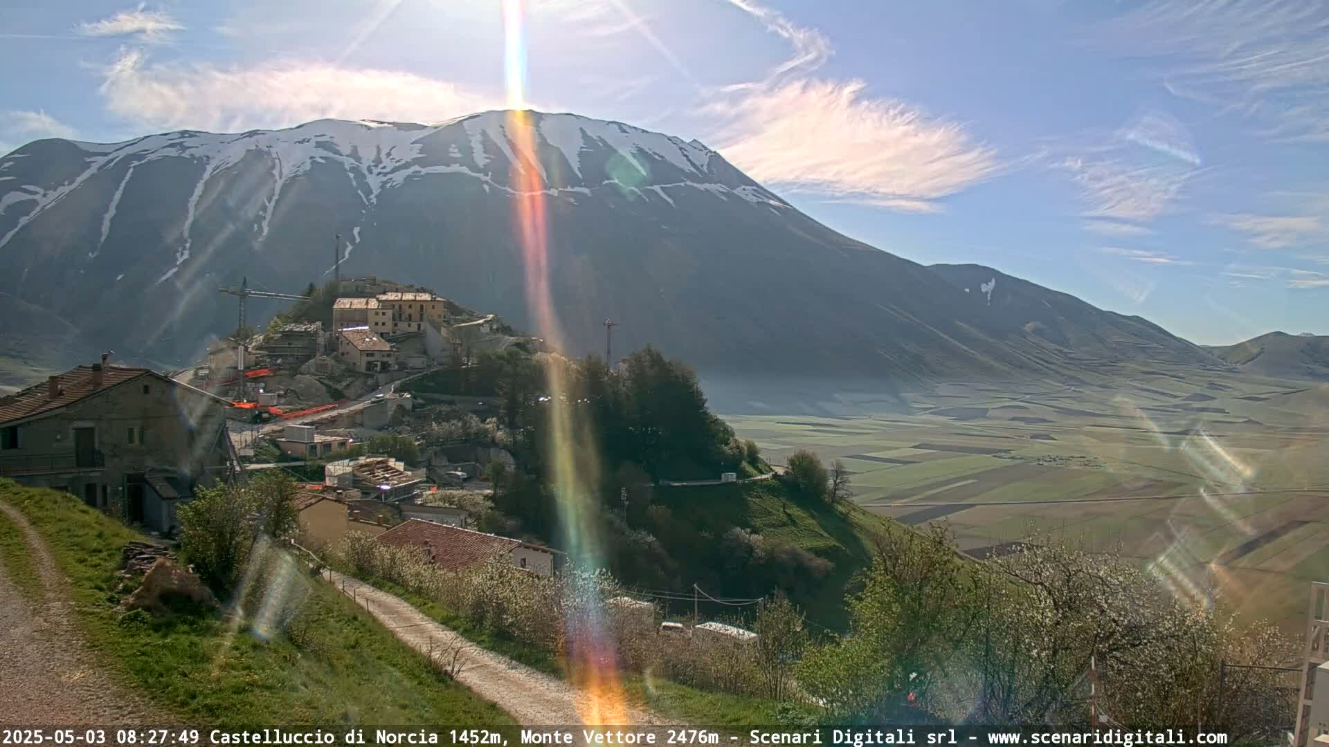 A sunlit mountain range with patches of snow overlooks a small village nestled in a valley of green and brown fields under a mostly clear sky.