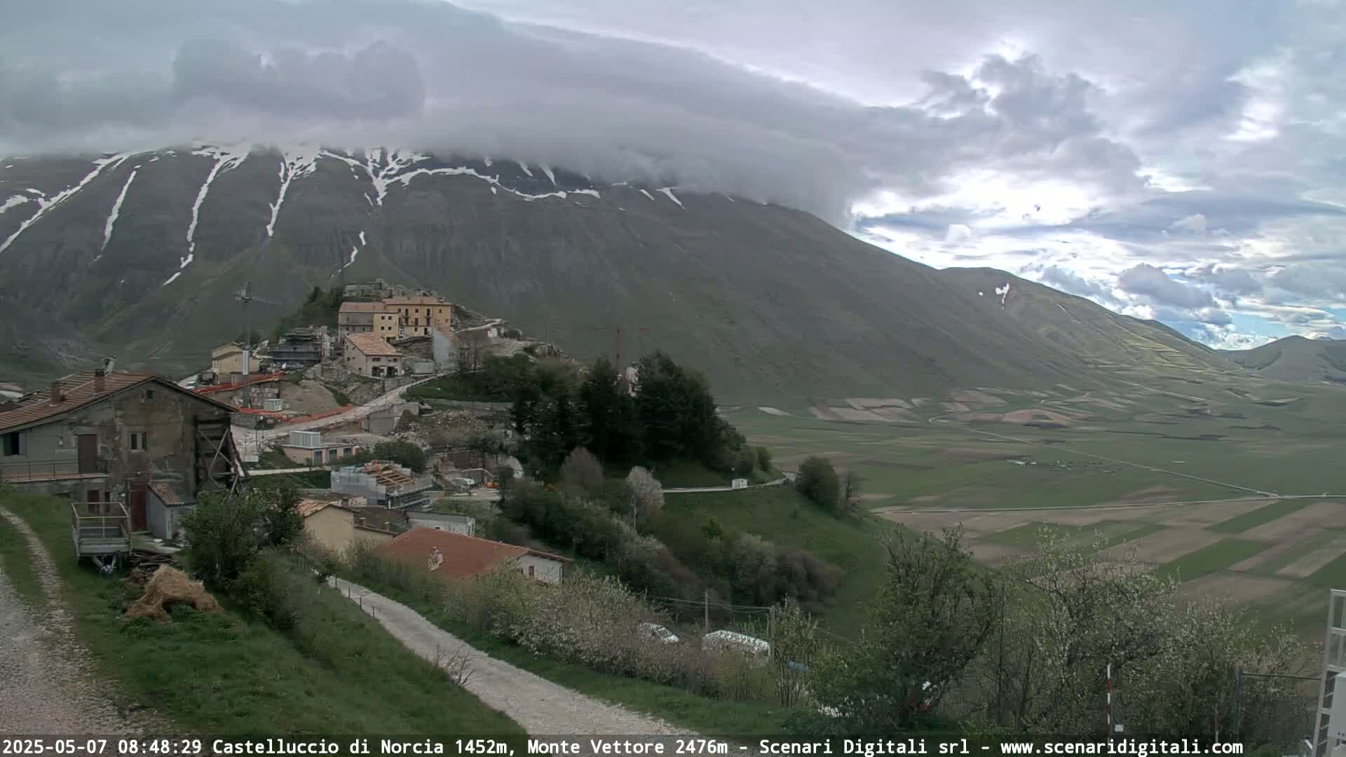 A mountain village nestled in a valley under a cloudy sky, with snow visible on the mountain peaks.